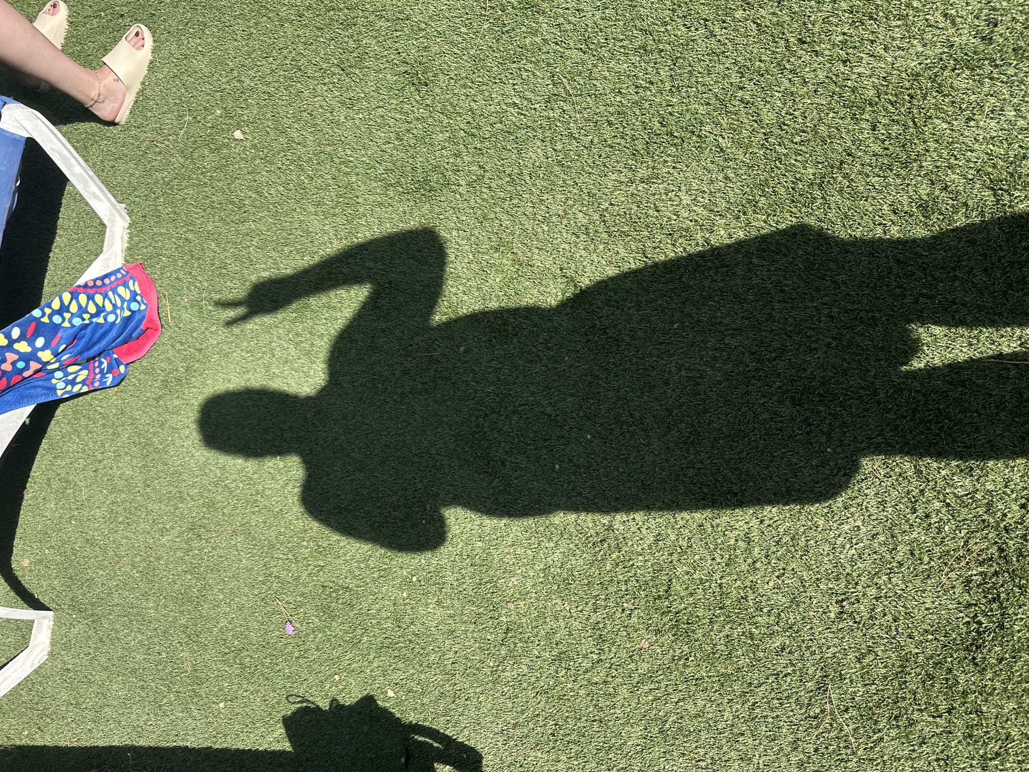 The image shows a shadow of a person making a peace sign on green artificial turf. The person's shadow is centered in the frame, with its head and upper torso towards the top of the image. The shadow's left arm is raised, with the index and middle fingers extended in a peace sign. The shadow's right arm is down. The turf appears to be in an outdoor setting, possibly a public space or a private yard. In the upper right portion of the image, a person's legs and feet are visible, wearing beige slides and a bracelet. A colorful towel or fabric is draped over a white lounge chair in the background, to the left of the person's legs. The lighting suggests it is daytime, with a strong light source casting the shadows. The location is identified as Sant Feliu de Guíxols, Spain.