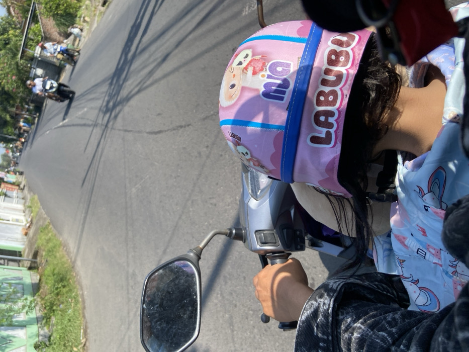 A person, likely a young girl, wearing a pink and blue helmet with a cartoon character and the words "Mia" and "Labubu" on it, is sitting on a scooter. The scooter is moving forward on a paved road in Mojosari, Indonesia. The girl is wearing a light blue shirt with cartoon unicorns on it. The road is empty in front of the scooter. The view is from the back of the scooter. There are houses on either side of the road, and green trees and vegetation lining the road. The sun is shining, and the weather appears to be sunny. Two motorbikes are visible in the distance.