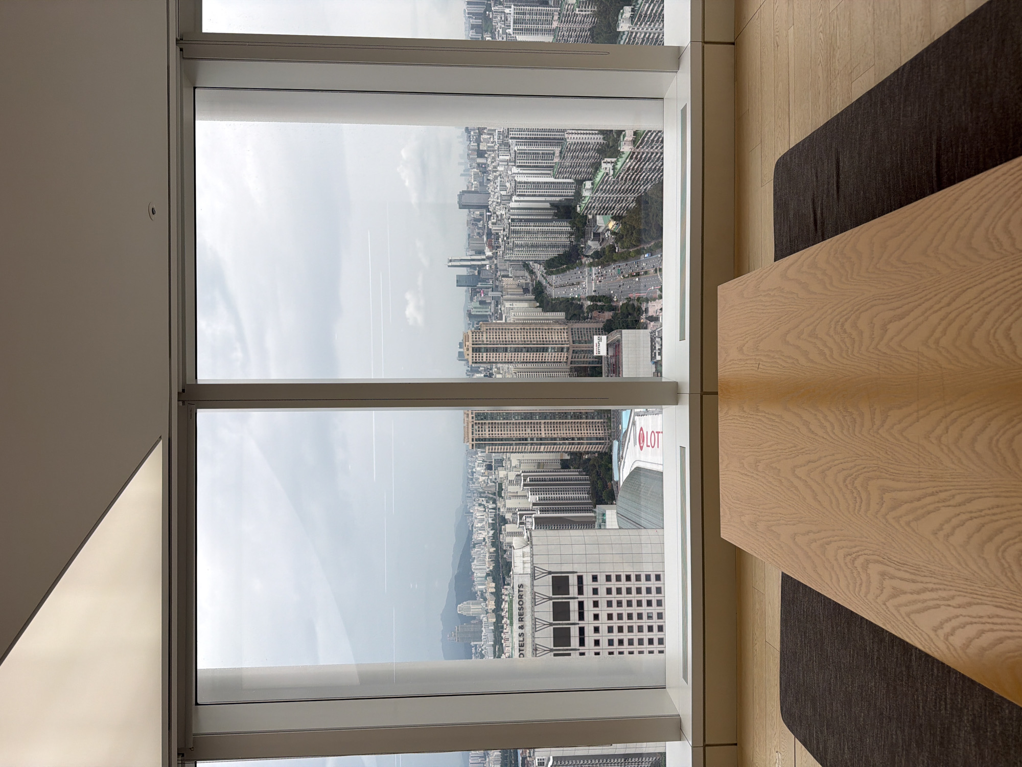 An interior shot captures a view of the cityscape of Seoul, South Korea, through a large window. In the foreground, a light-colored wooden table is positioned on a dark rug, which is on a light-colored wooden floor. The table is angled towards the right side of the frame. Beyond the table and rug, the expansive window reveals a dense urban landscape under an overcast sky. Numerous high-rise buildings of varying heights and architectural styles dominate the view, interspersed with roads and greenery. Notable structures include a building with the text "HOTELS & RESORTS" visible, and another complex with "LOTTE" signage. The cityscape extends to the horizon where mountains are faintly visible. The scene suggests a moment of quiet observation from a high vantage point.