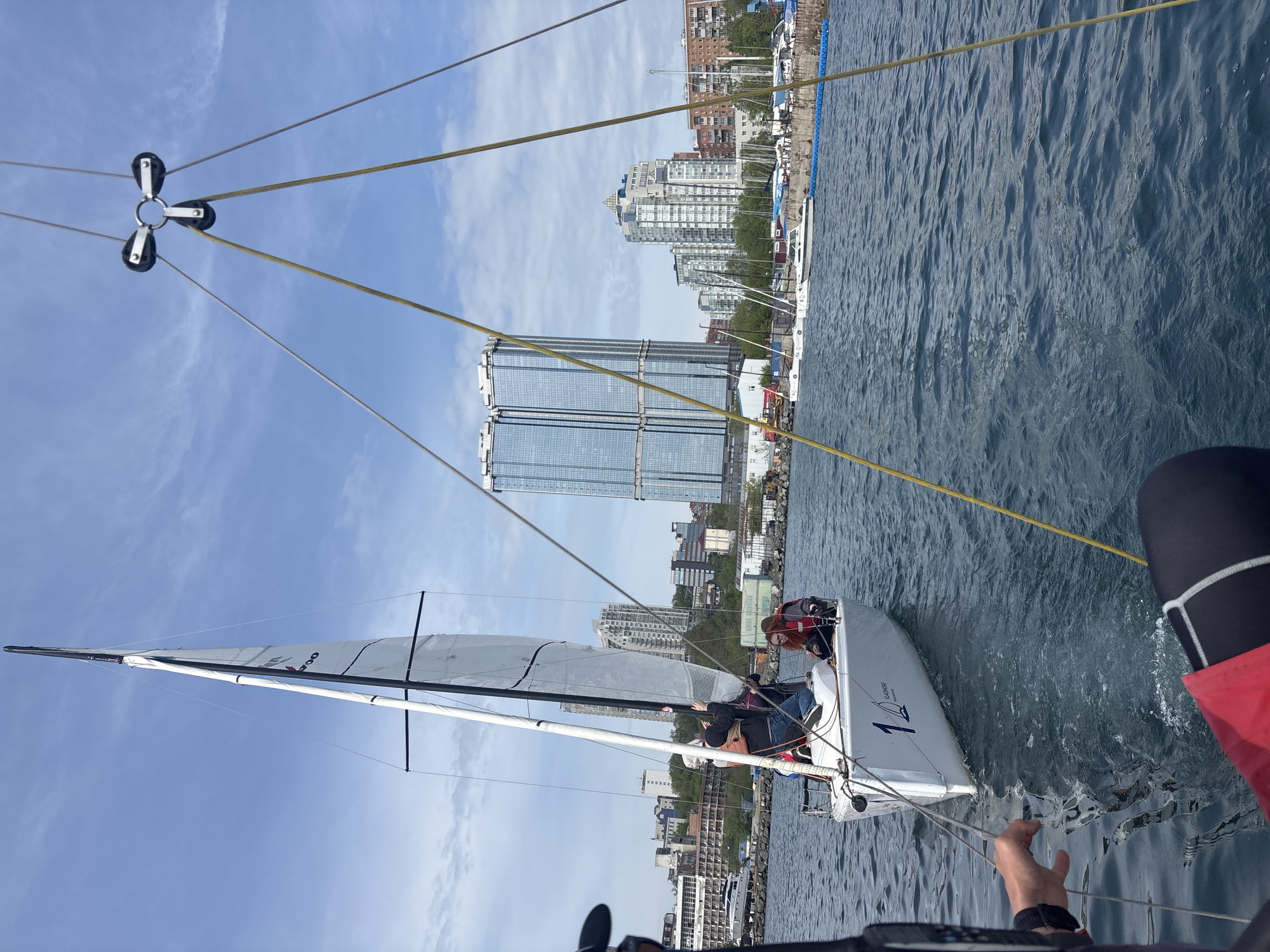 Here is a detailed description of the image:
The image is a slightly high-angle shot taken from what appears to be another boat, focusing on a white sailboat in a body of water near an urban setting. The sailboat has one sail hoisted. There are two people on the sailboat, one of whom is wearing a red life vest.
The sailboat appears to be rigged with lines and pulleys, one of which is visible in the top portion of the frame. The lines extend from the sailboat upwards towards a system of pulleys. The water around the boat is choppy, indicating slight wind activity.
In the background, there is a city skyline featuring tall buildings and structures. The buildings suggest an urban environment with a mix of residential and commercial structures. The city skyline is identifiable as Vladivostok, Russia, due to the presence of distinctive buildings, including the two tall, matching buildings with mirrored glass facades. A few boats are docked near the coastline behind the sailboat. The sky is mostly clear with a few scattered clouds.