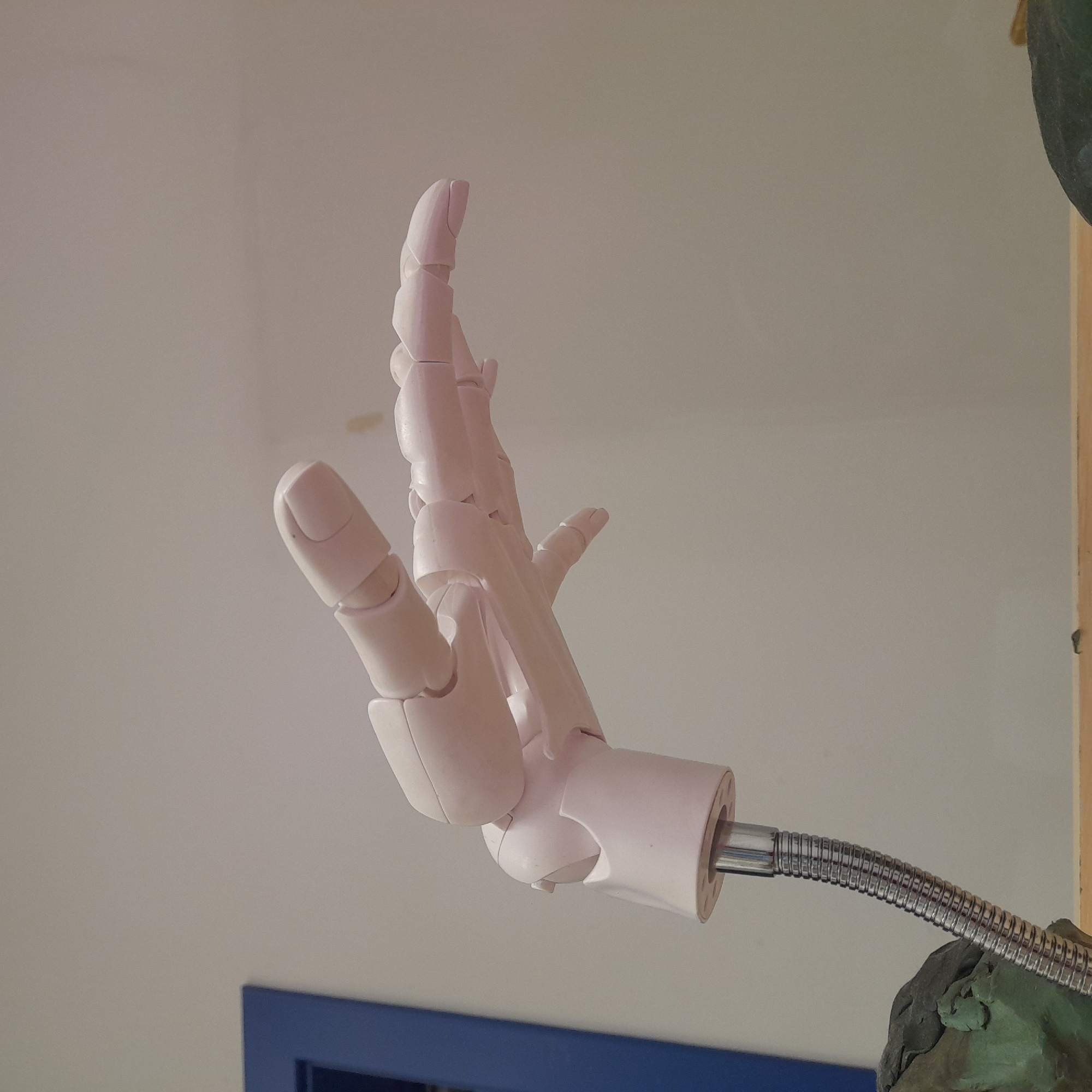 A close-up, eye-level, medium shot shows a pale pink, articulated robotic hand reaching outwards and upwards. The hand is attached to a flexible, metallic silver gooseneck tube which is supported by a textured, dark green clay-like material. To the left of the image, a vertical blue door frame is partially visible. The background consists of a plain, light grey wall. The lighting is even and neutral, casting no distinct shadows. The image does not provide information about the location in the city.