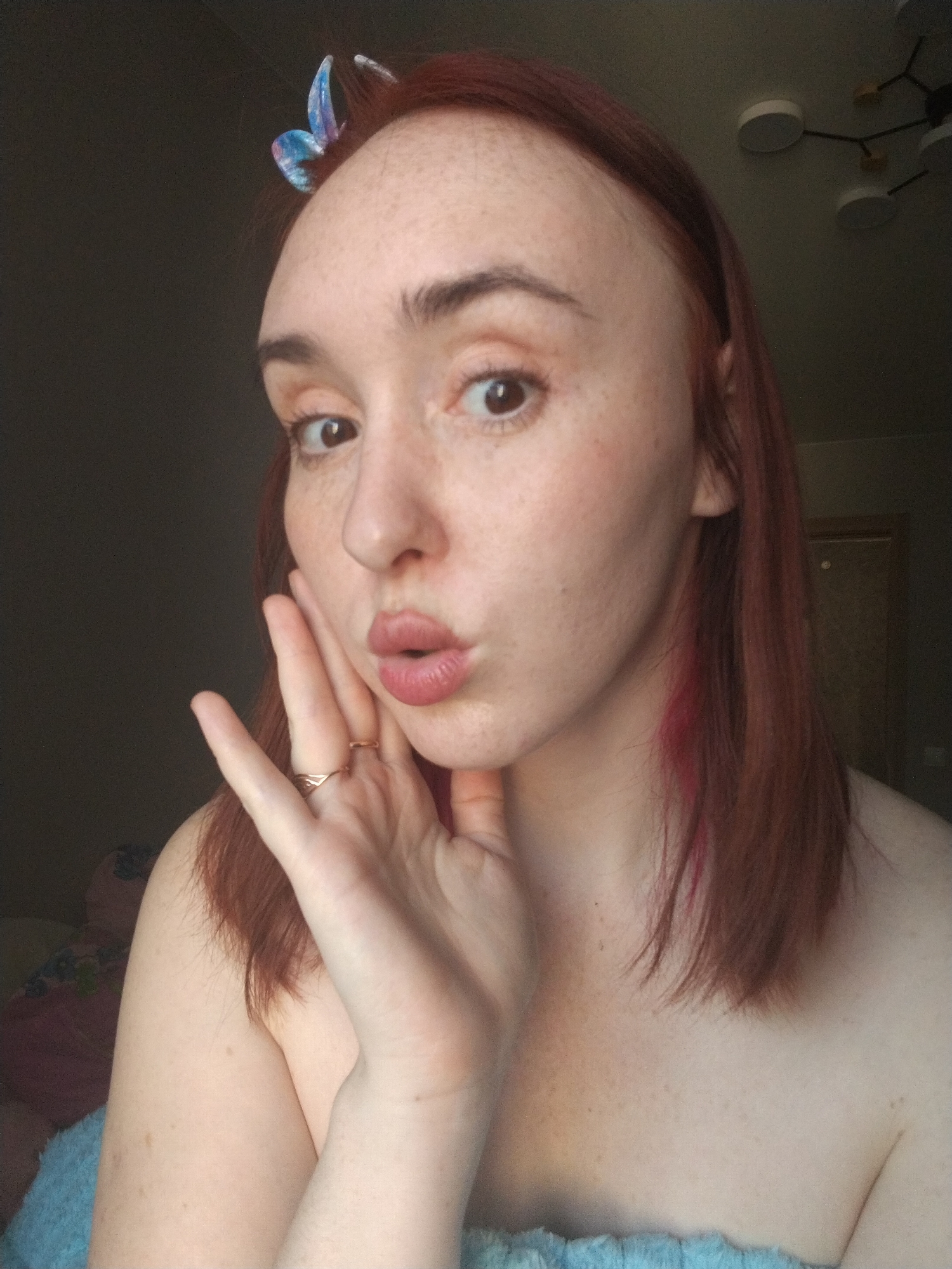 A woman with red hair, freckles, and a butterfly clip is posing for a selfie indoors. She is wearing a light blue towel or garment around her shoulders. Her hand is placed on her cheek, and she is puckering her lips. There is a ring on one of her fingers. The background is somewhat dark, but a modern light fixture can be seen on the ceiling. A doorway is partially visible in the background. The scene is in a room with a bed, with a plush toy on it. There is no direct evidence to confirm the location in Izhevsk, Russia.
