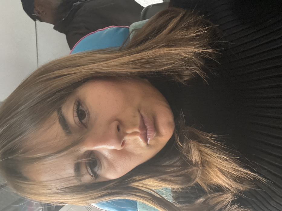 The image displays a close-up, front-facing portrait of an adult female, likely a selfie, taken indoors. The woman has medium brown hair with bangs styled across her forehead and falling to her shoulders. Her eyes are dark, and her lips are pursed in an expression resembling a "kissing" or "duck face." She is wearing a black, ribbed turtleneck sweater.
In the blurred background, to the left of the woman's head, a light blue seat headrest or cushion with a distinct pink border is visible. To the right, a dark-skinned male is partially visible, out of focus, wearing a dark jacket and a dark cap or hat. The environment suggests the interior of a vehicle, possibly a train, bus, or airplane, due to the visible seating and general cabin appearance.
The woman is directly engaged with the camera. There is no discernible interaction between the woman and the individual in the background. The specific location within the city of Lançon-Provence, France, cannot be determined from the visual information presented in the image.