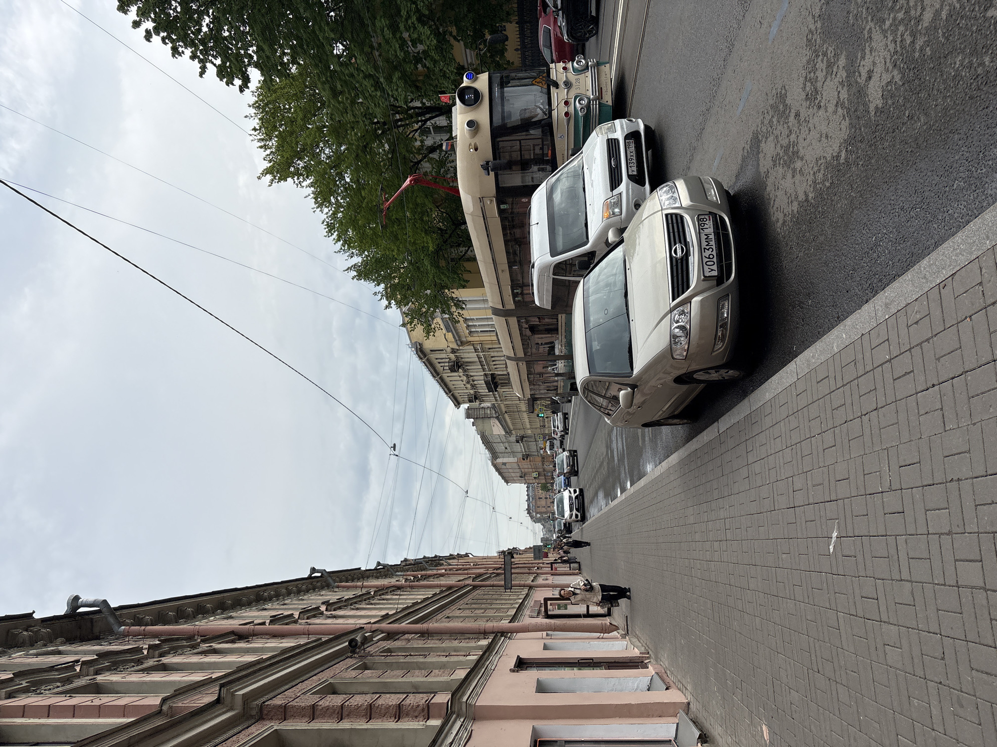 The image captures a street scene in Saint Petersburg, Russia. The street is lined with buildings, a sidewalk made of grey brick, and parked cars along the curb. A tram is partially visible in the background and is also parked. Overhead electrical lines crisscross the street. A person is seen standing on the sidewalk near the bottom right of the frame. The weather appears overcast.