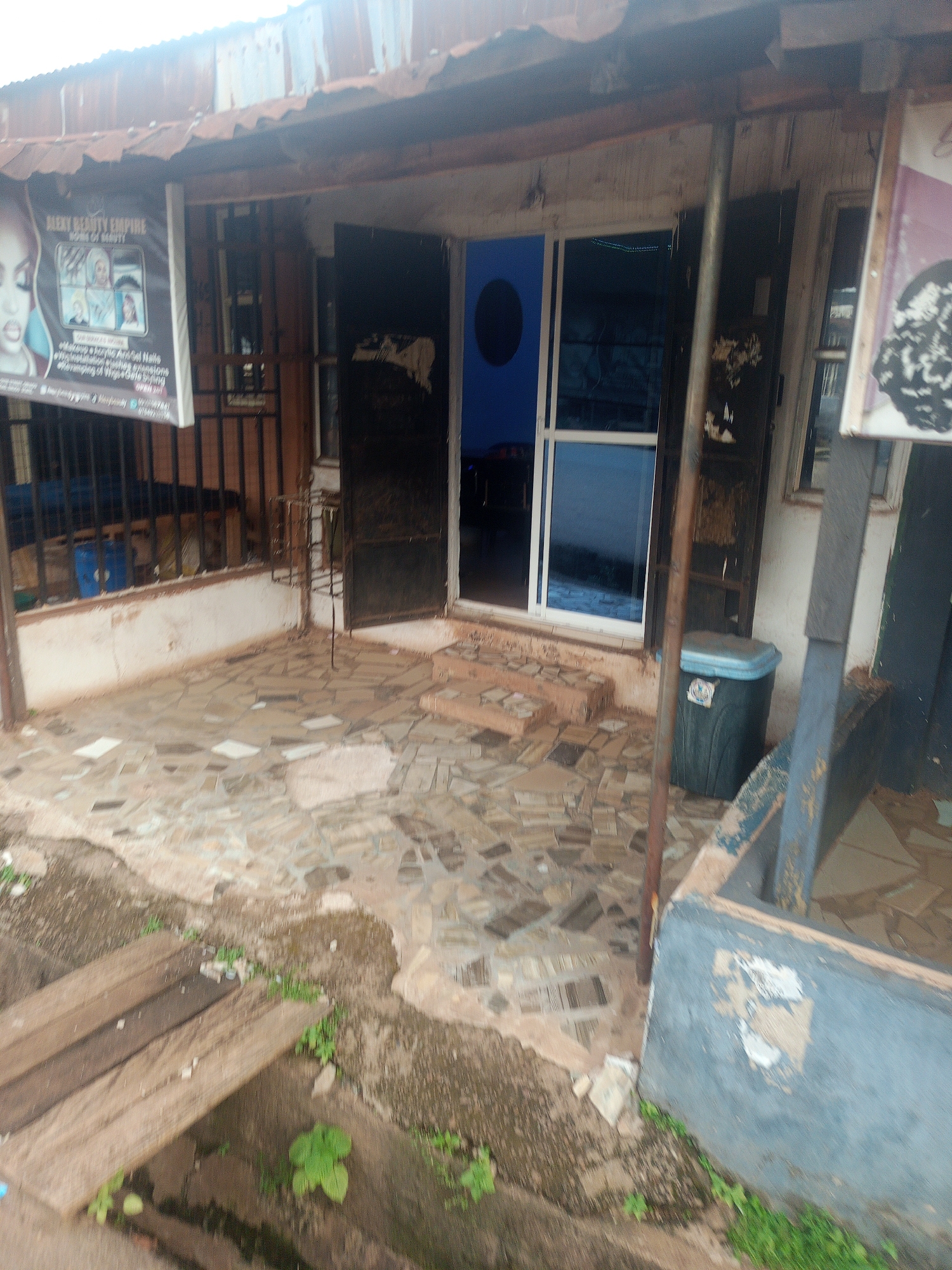 The image depicts a storefront in Enugu, Nigeria, that appears to be a beauty salon or salon. The facade of the building is constructed from corrugated metal roofing and has an aged, somewhat dilapidated appearance. A sign advertising "ALEXY BEAUTY EMPIRE" is prominently displayed on the left side of the entrance. This sign features images of women's faces and text detailing services such as "Makeup & Acrylic And Gel Nails," "Facial Treatment," and "Hair Extension."
The entrance consists of two large doors, one of which is open, revealing a blue-tinted glass interior. The ground in front of the shop is paved with irregularly shaped stones. To the right of the entrance, a dark green trash bin with a light blue lid stands near the wall. The overall environment suggests an outdoor commercial space in an urban setting, with evidence of wear and tear on the structure and surrounding area. Green vegetation is visible growing around the base of the building and in cracks in the pavement. A wooden plank walkway is present in the foreground, indicating a possible drainage or elevated path.