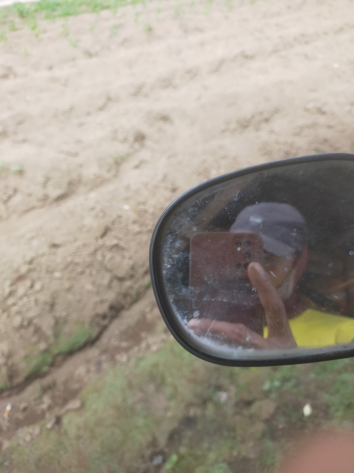 The image shows a close-up of a dusty side-view mirror, reflecting a person taking a selfie with a smartphone. The person is wearing a dark baseball cap and a yellow shirt. The background of the mirror shows a field of dry earth with some sparse green vegetation. The overall setting suggests an outdoor environment, possibly rural, given the dry earth and minimal vegetation. There is no visible indication of the specific location within Padang Dalom, Indonesia, as the image lacks any identifiable landmarks or contextual clues.