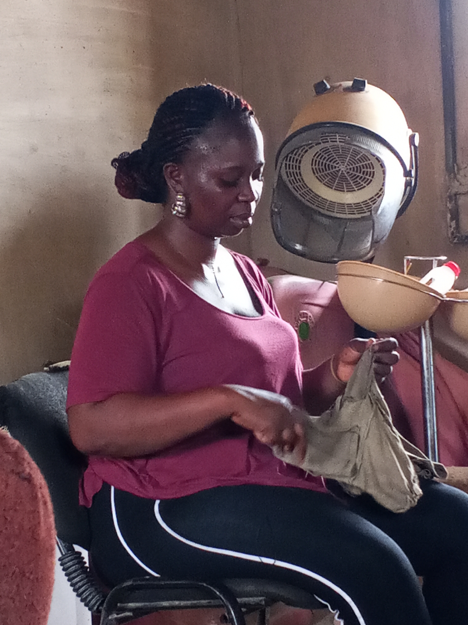 A woman sits in a chair in a beauty salon in Adonkolo, Nigeria. She is wearing a maroon t-shirt, black leggings with a white stripe, and dangling earrings. Her hair is styled in braids pulled back into a bun. She is holding and examining a tan cloth. Behind her is a salon hair dryer on a stand. A counter with bowls and bottles is to the right. The walls of the salon are tan.