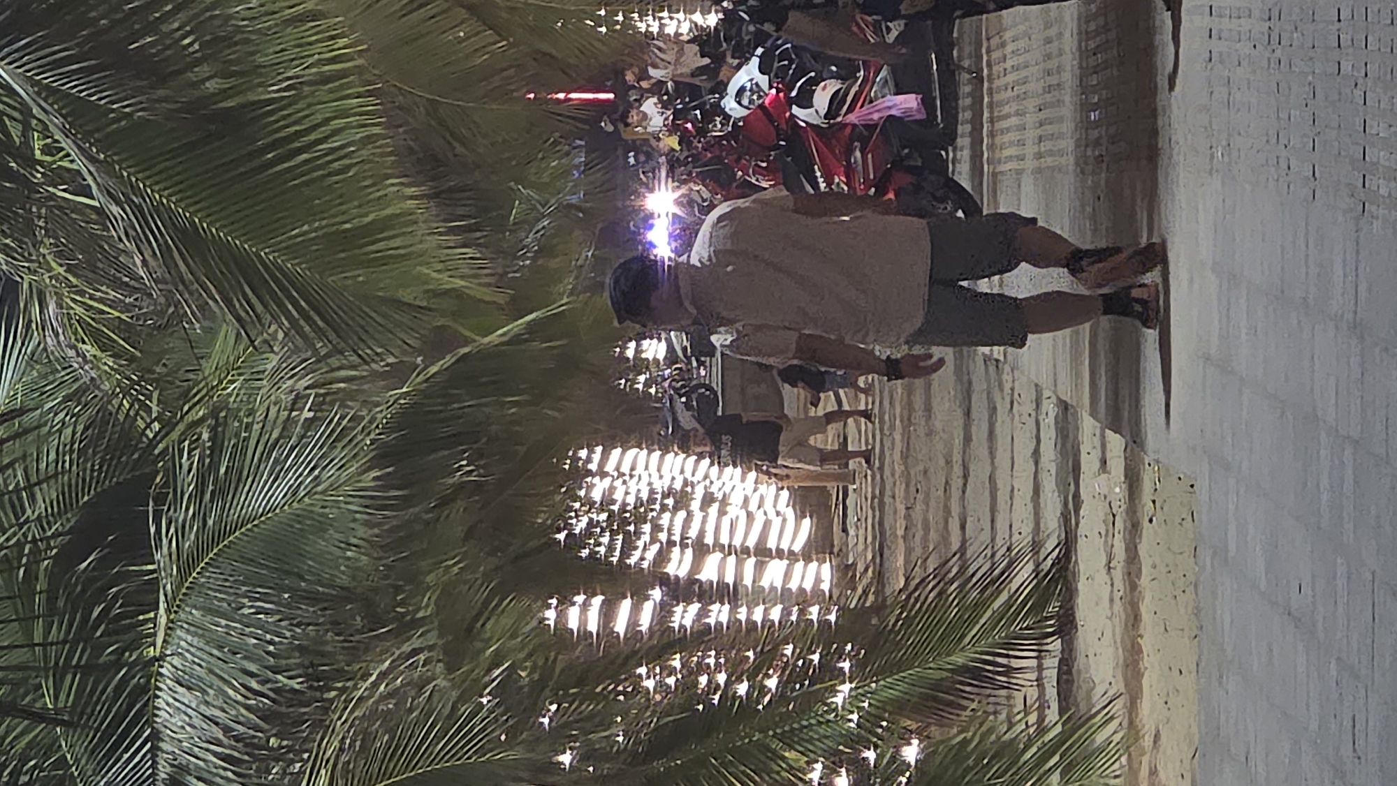 A nighttime image depicts a scene along a street in Nha Trang, Vietnam. A man in a light-colored shirt and shorts walks on a pavement along a busy street with motorbikes. Palm trees line the walkway. The street is illuminated by lights, reflecting on the pavement. Crowds of people are gathered near the motorbikes. The scene appears lively and busy.