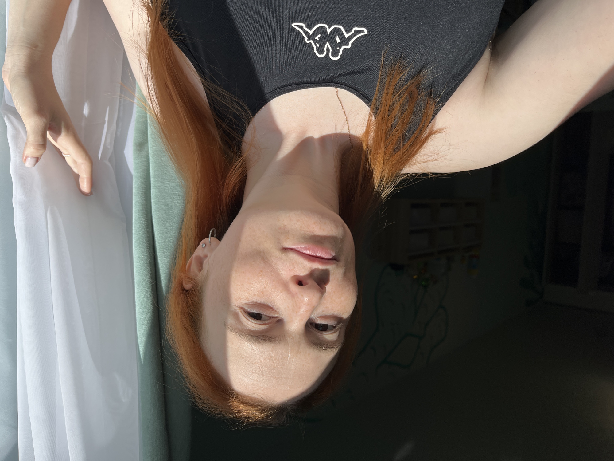 An outdoor, close-up, head-and-shoulders shot shows a woman with fair skin and freckles, and long, reddish-orange hair. She is wearing a black tank top with a white Kappa logo on the chest, and a thin gold chain. Her left arm is bent, and her hand is resting on her bicep. Her right hand is reaching towards a sheer, white curtain that hangs to her right. The lighting suggests it is daytime, with sunlight casting shadows and highlighting her face. The background is dimly lit, with what appears to be a light-green wall adorned with a teal drawing and shelves in the distance. The image was taken in the city of Chaikovsky, Russia.