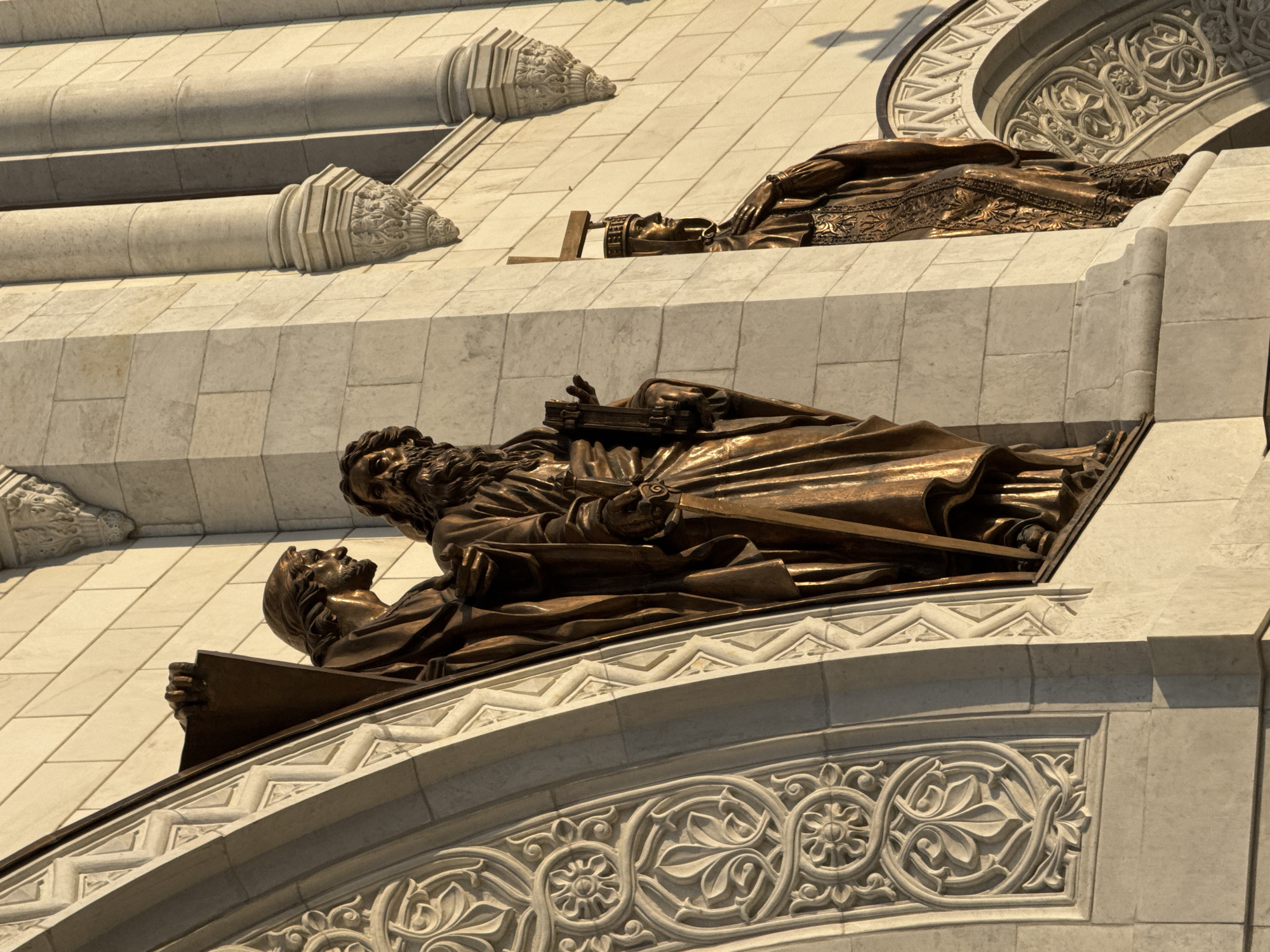The image shows a close-up of bronze sculptures on the exterior of the Cathedral of Christ the Savior in Moscow, Russia. Two bronze figures, possibly depicting biblical figures, are lying on ledges or platforms. The lower sculpture shows one bearded man laying on top of a man with a book. The architecture includes decorative stone elements with carved details. The building appears to be made of light-colored stone or brick.