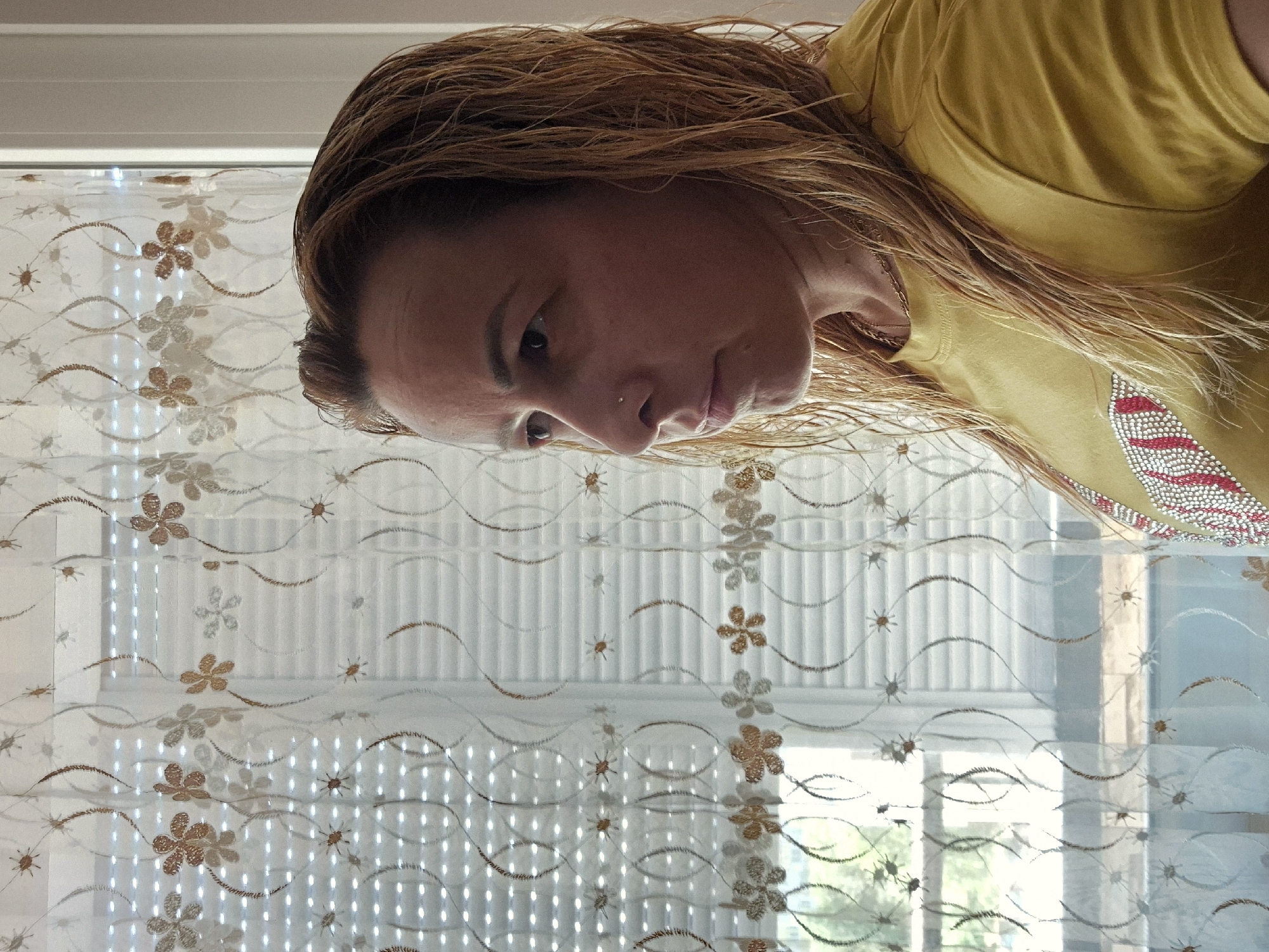 A medium shot shows a woman with wet, light brown hair and a nose ring looking to the left of the frame. She is wearing a yellow t-shirt with a design of red and white stripes outlined by rhinestones. The woman is standing in front of a sheer white curtain with a floral pattern in brown and gold. Behind the curtain, blinds are visible, and beyond that, a window shows greenery and what appears to be a building. The lighting suggests it is daytime. The setting is indoors.