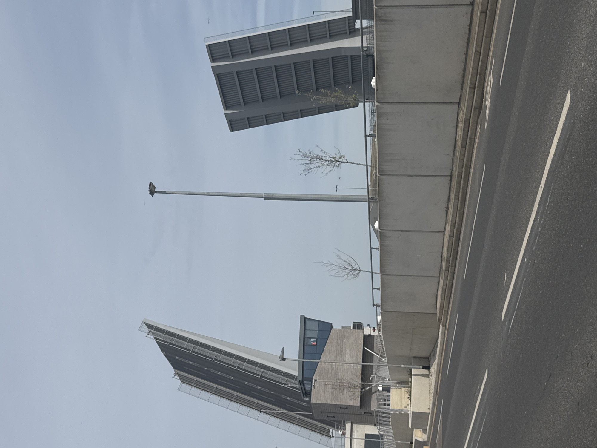 The image depicts a partially opened drawbridge located in Gorleston-on-Sea, United Kingdom. The bridge is constructed of gray concrete and metal, featuring two sections raised at angles to allow passage for marine traffic. A street, marked with white lines, runs alongside the bridge’s infrastructure. Several bare trees are visible along the bridge's length. A tall lamppost stands near one section of the bridge. A control tower-like structure is situated between the raised bridge sections. The sky is overcast.