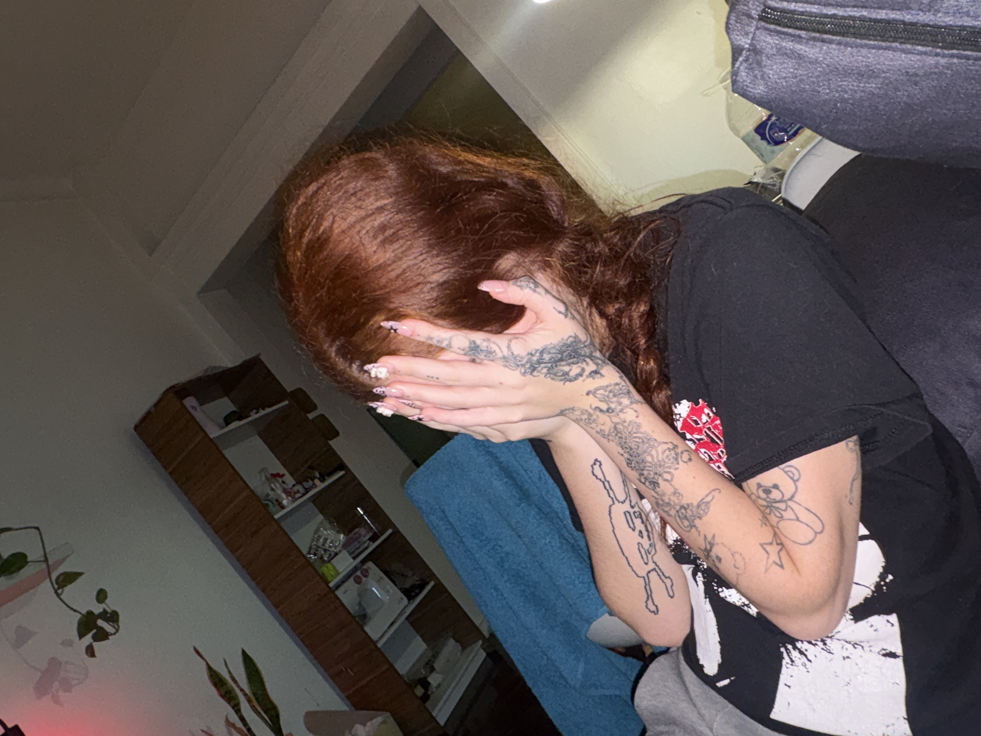 A person with reddish-brown hair is captured covering their face with both hands, their head bowed downwards. They are wearing a black t-shirt featuring a white splattered graphic design with red elements, and grey bottoms, likely sweatpants. Their hands and forearms are extensively covered in tattoos, showcasing various designs including a teddy bear, a star, and other intricate, abstract patterns. Long, decorated nails are visible on their fingers.
The scene is an indoor setting, illuminated by dim, artificial lighting, suggesting it is evening or night. The person appears to be seated or reclining, with a textured blue blanket draped over part of their body. In the background, a tall, wooden bookcase or shelving unit holds various objects, including bottles and containers; a sewing machine can be discerned on a lower shelf. Potted plants, including a trailing vine (resembling a pothos) on a wall shelf and a snake plant on the floor, add to the decor. To the upper right, a dark blue zippered bag or cushion is partially visible. Behind the person's shoulder, a white bottle with a blue and white label, bearing indistinct text, can be seen.
The pose of covering the face conveys a sense of shyness, introspection, or a desire for privacy. The image provides a glimpse into a personal moment within what appears to be a cozy indoor space in Buenos Aires, Argentina.
