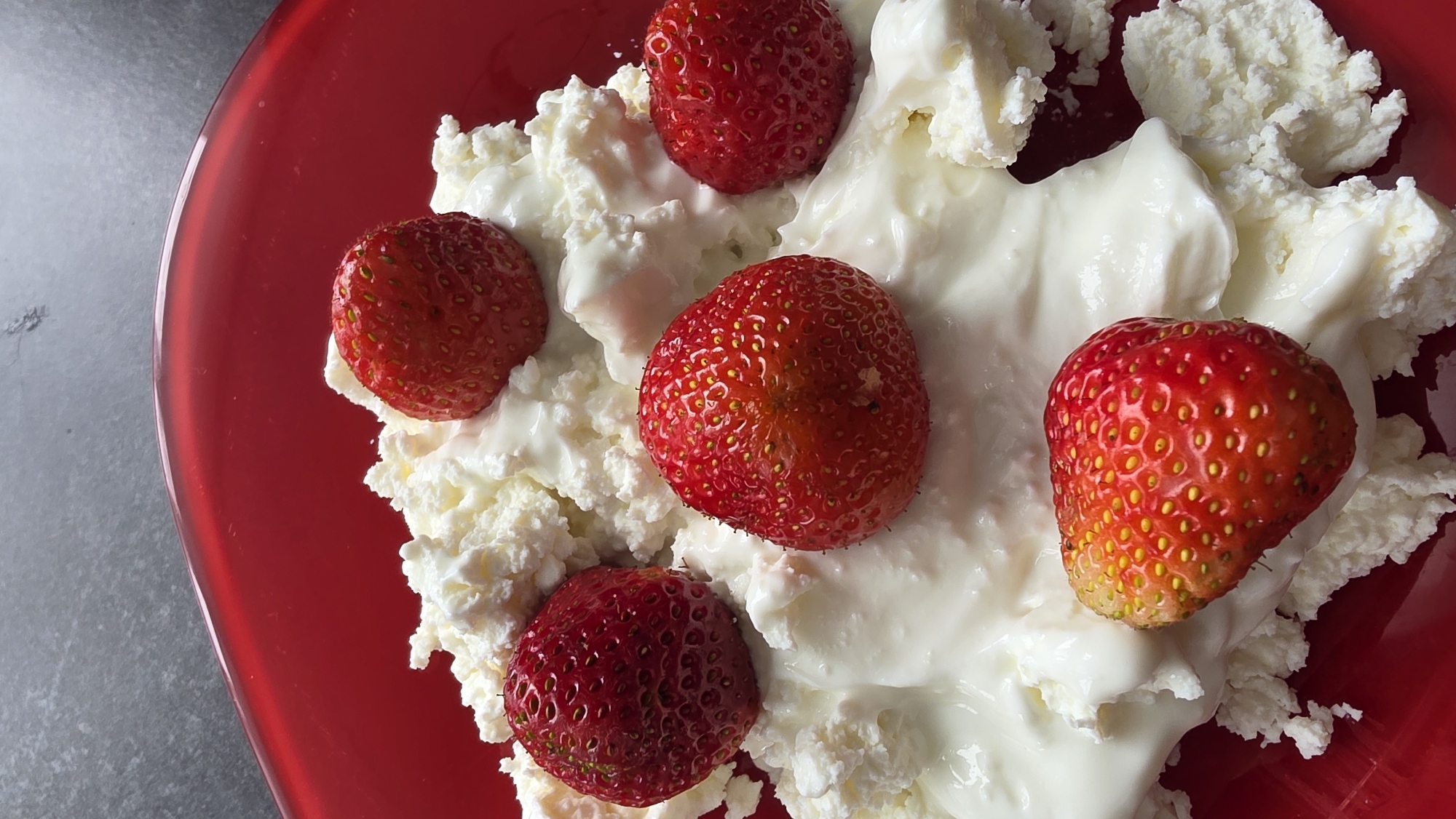 A close-up, overhead shot shows a red bowl filled with cottage cheese and topped with five strawberries. The cottage cheese is lumpy and white, with dollops of a smoother, yogurt-like substance mixed in. The strawberries are a vibrant red, with visible seeds and green stems on some. The background is a dark gray, textured surface.