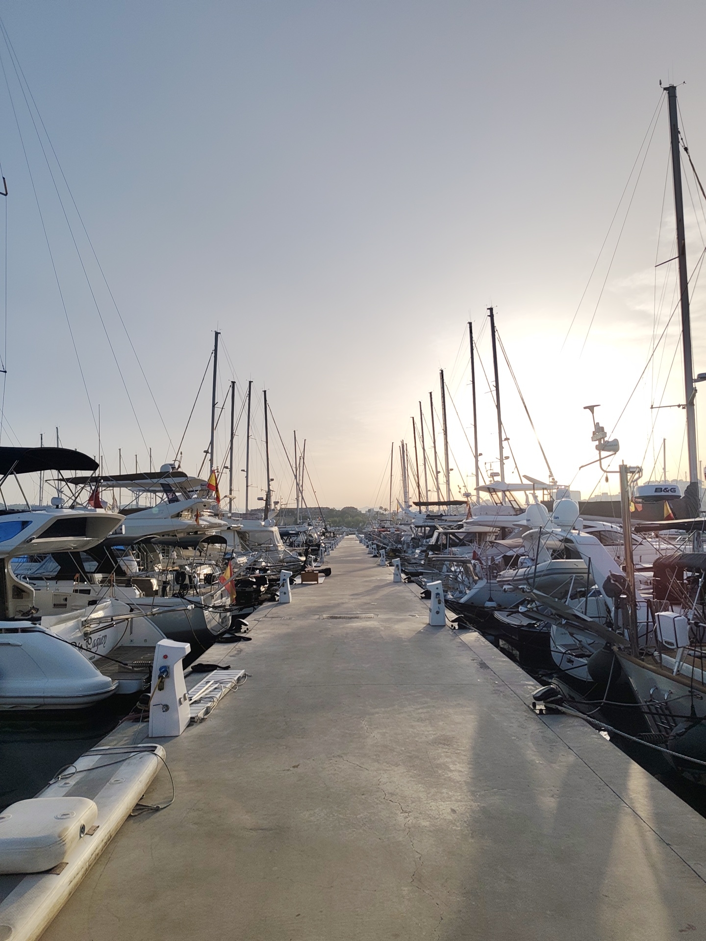 The image depicts a marina in Dénia, Spain, at sunset or sunrise. A concrete pier runs down the center of the frame, flanked by rows of moored yachts and sailboats. Masts rise high above the vessels, creating a forest of vertical lines against the bright sky. Spanish flags are visible on some of the boats. The pier is bare, showing no immediate signs of human presence.