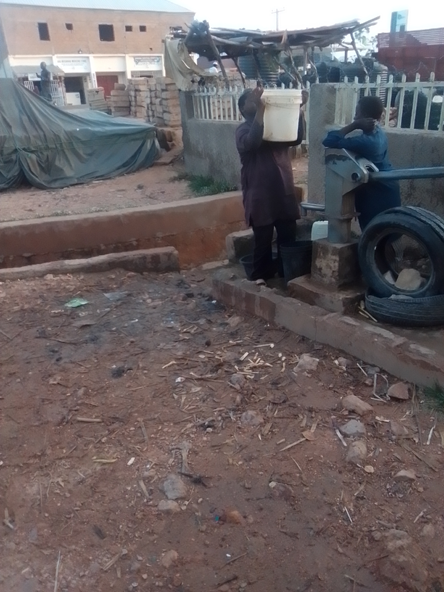 In Pantame, Nigeria, a young boy in a blue shirt stands near a manual water pump. He is reaching out and appears to be interacting with the pump mechanism. To his left, another person, possibly an adult, in a dark maroon garment is holding a white bucket above their head, as if to collect water. The ground is dirt and debris, with a concrete structure and some discarded tires visible. In the background, a building with a light-colored facade and multiple windows suggests a commercial or residential area. A tarp-covered structure and stacks of bricks are also present, indicating ongoing construction or a marketplace. The overall environment appears somewhat unkempt, with scattered litter and organic matter on the ground.