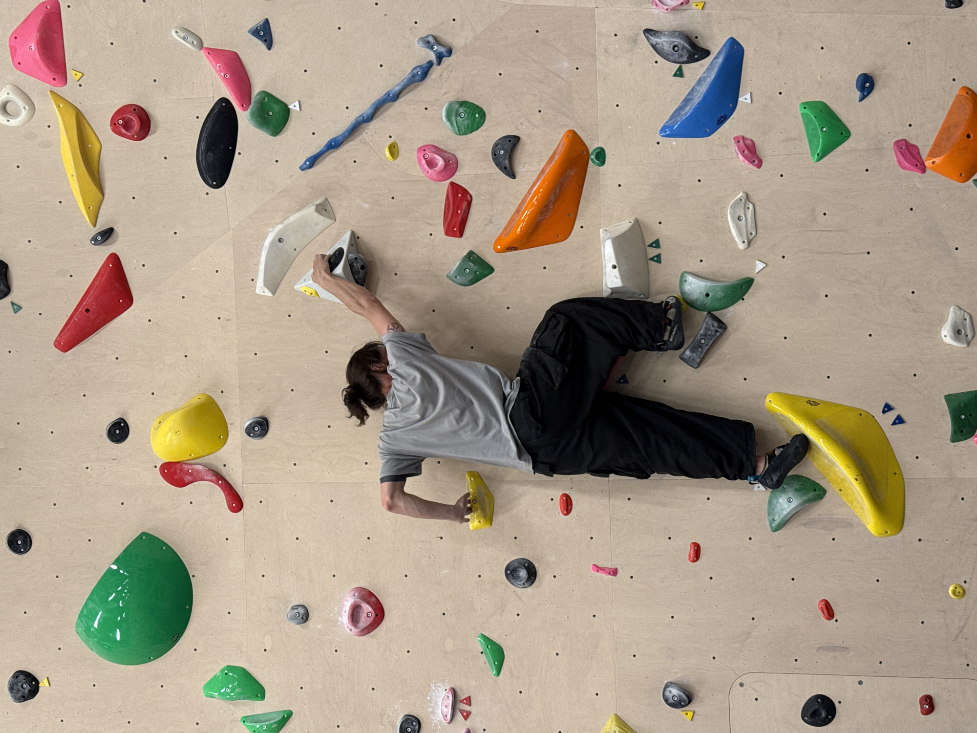 A person is suspended on a climbing wall, gripping holds with hands and feet. The wall is constructed of plywood, and various colorful climbing holds are bolted to its surface. The person, dressed in a grey shirt and black pants, is horizontally oriented. There is no visible indication of the city or location as Sochi, Russia.