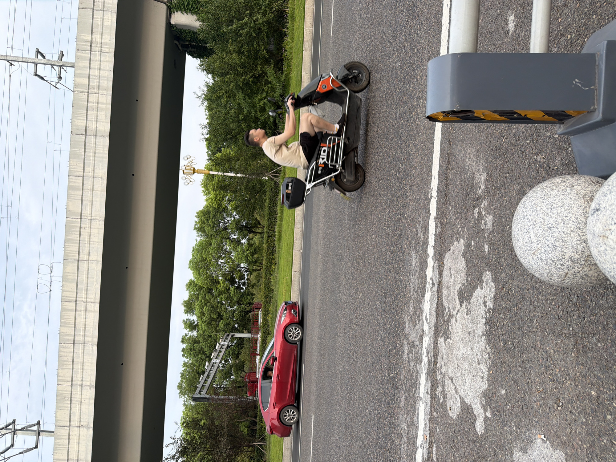 In Jiangjin, China, a man rides an electric scooter on a paved road under an elevated highway. The scooter is black and orange with a black storage box on the back. The man wears a light-colored t-shirt and dark shorts. To his left, a red sedan is driving in the opposite direction. The sedan is a Mazda and has tinted windows. In the background, there are trees, a lamppost with multiple lights, and overhead power lines. The road has white lane markings. In the foreground, there are two large, spherical granite bollards and a section of a grey barrier with yellow and black hazard stripes. The sky is overcast.