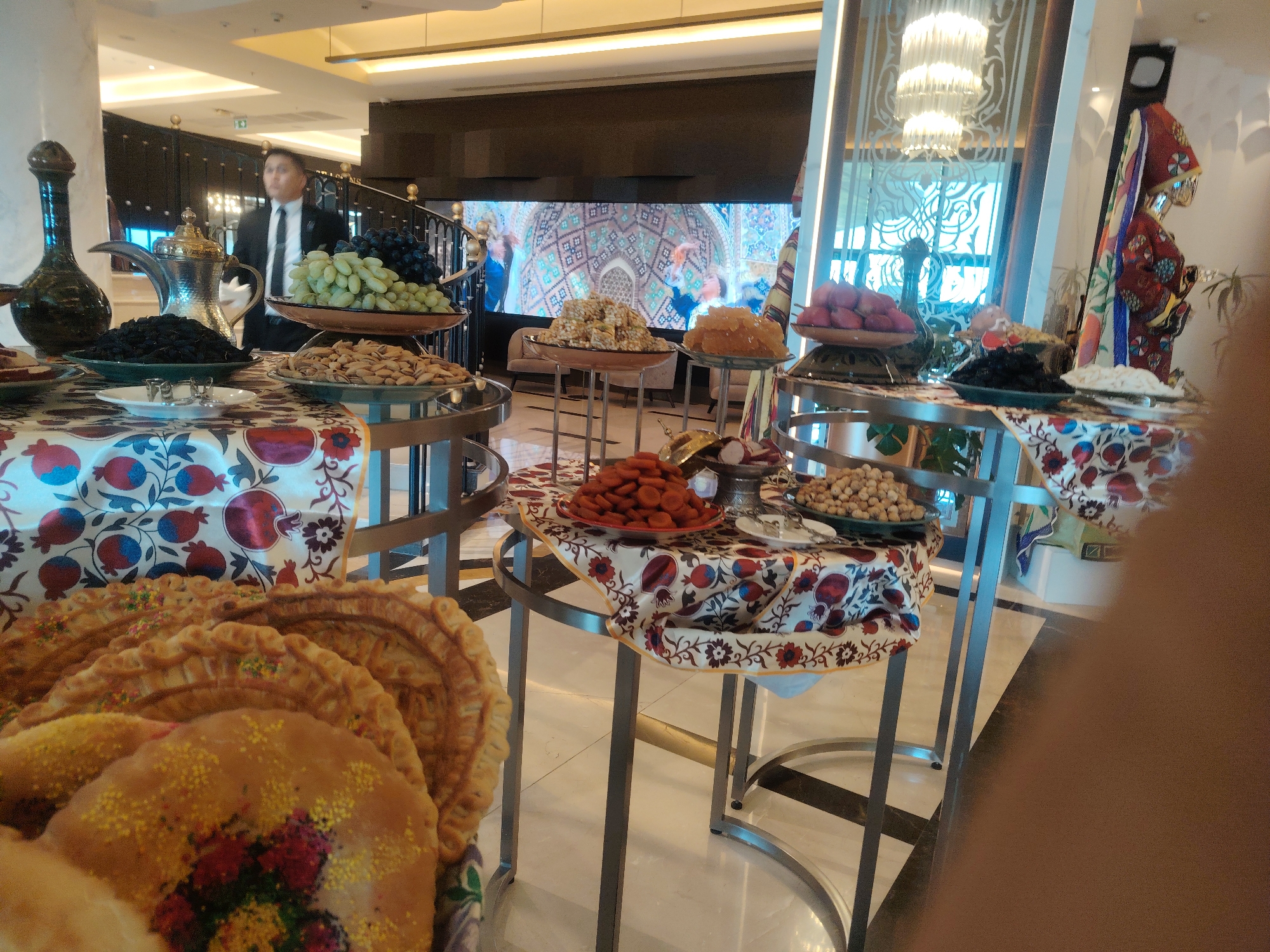 The image shows a buffet setup with various food items, likely in Konigil, Uzbekistan. In the foreground, there are several round, decorated pastries, possibly samsa or pies, arranged on a platter. Behind these pastries, a table is covered with a fabric featuring a pomegranate and floral pattern, displaying bowls of dried fruits and nuts, including pistachios and what appear to be dried apricots. Further back, another table holds more fruits like grapes and dried dark berries, along with a traditional teapot and a decorative jug. A man in a suit and tie stands behind the buffet, appearing to be a staff member. In the background, a large screen displays an intricate mosaic-like pattern, and to the right, a mannequin dressed in traditional Uzbek attire is visible. The environment suggests an indoor setting, possibly a hotel lobby or a reception area, with polished floors and modern lighting fixtures.