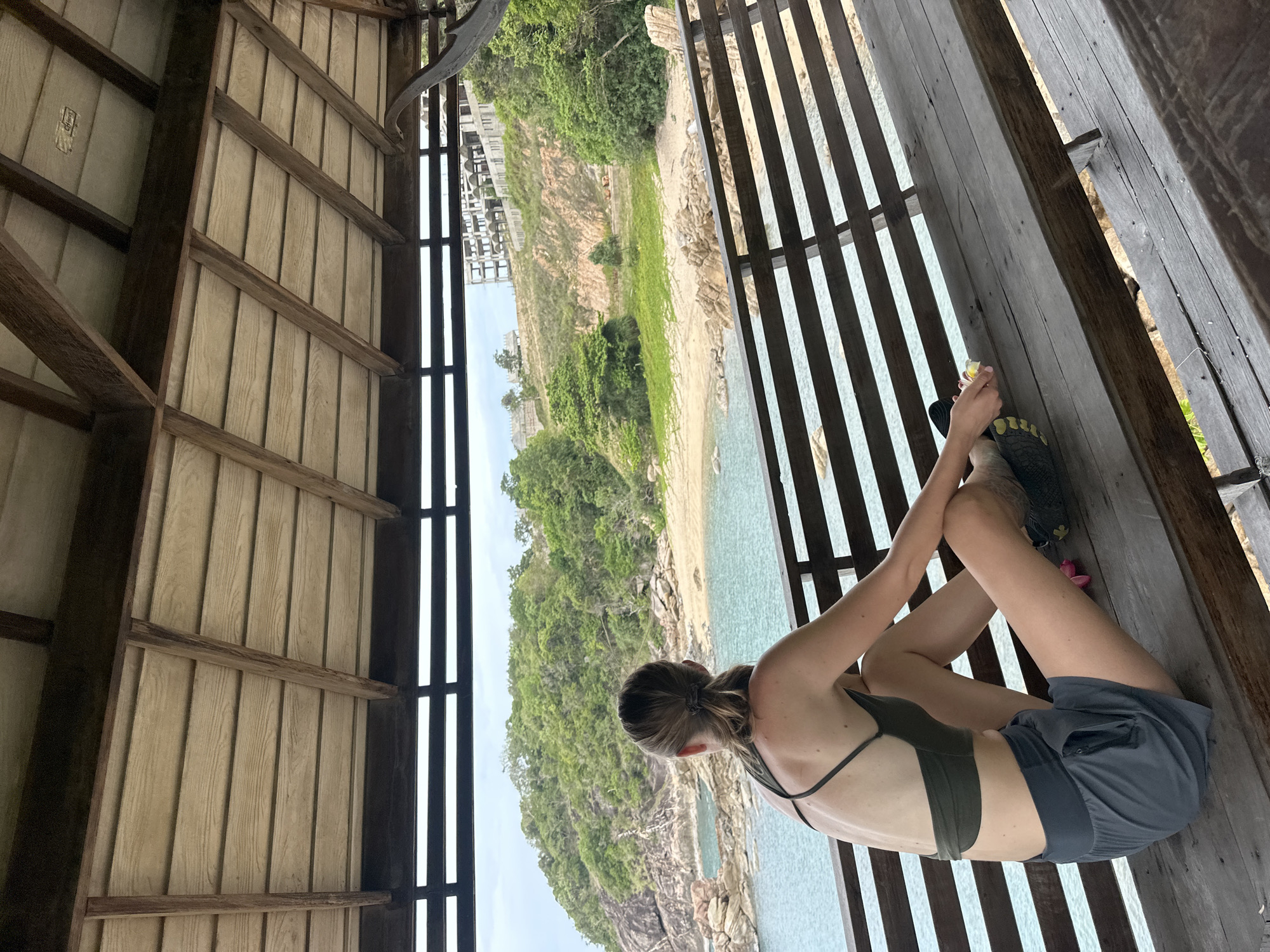 A young woman with her hair in a ponytail, wearing a dark green sports bra and grey shorts, sits on a wooden bench under a thatched roof structure. She is looking out at a scenic view of a rocky coastline, turquoise water, and a lush green hillside dotted with buildings. The bench is made of weathered wooden planks, and the roof is constructed of similar material supported by wooden beams. The woman's feet are bare, and she is holding a small pink flower in her right hand. The overall setting appears to be a relaxed, outdoor location, possibly a resort or a viewpoint on the island of Ko Samui District, Thailand.