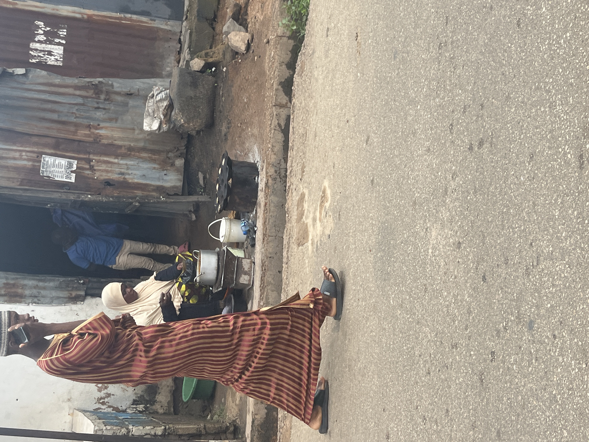 The image shows a street scene in Jos, Nigeria. In the foreground, a person wearing a striped robe and a patterned cap is holding a phone. To the left, a woman in a cream-colored headscarf is seated, also holding something in her hand. Behind them, a man in a blue shirt and light-colored pants is standing near an open doorway, which appears to be a shop or dwelling made of corrugated metal. Various objects, including pots and buckets, are visible in front of the structure. The ground is paved with asphalt, and there is a dirt and rocky area next to the road. The lighting suggests it is daytime. The overall scene depicts a casual street interaction in an urban environment. A notice with some visible text is affixed to the corrugated metal wall.