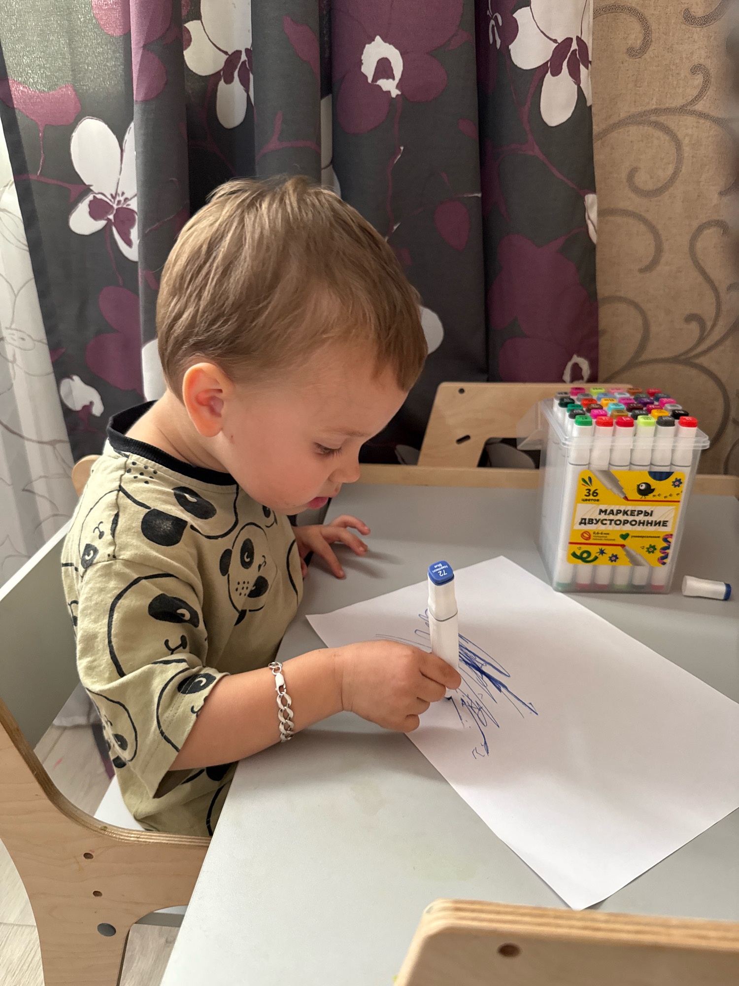 A young child with light brown hair is sitting at a grey table, drawing on a white piece of paper with a blue marker. The child is wearing a light tan t-shirt with a repeating pattern of panda faces. On the table, to the right of the child, is a clear plastic container filled with an assortment of colorful markers. The background features a grey curtain with a floral pattern of white and purple flowers, and a textured beige wall with a swirling pattern. The image appears to have been taken indoors, likely in a residential setting.