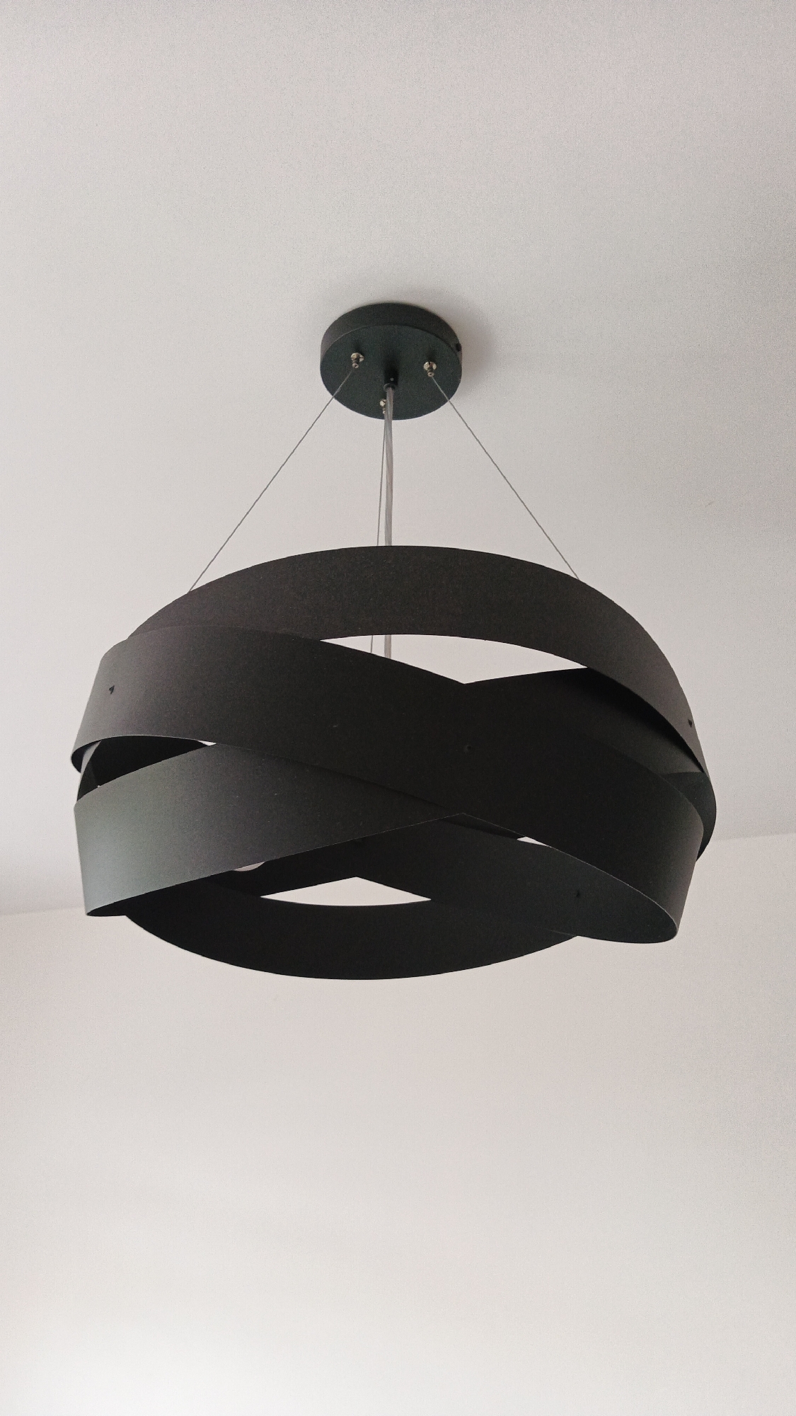 A black, modern chandelier with multiple overlapping ribbon-like elements hangs from a white ceiling. The chandelier is suspended by thin wires from a circular black canopy. The background is a plain white wall and ceiling, typical of an interior residential or commercial space. There are no people or other objects visible. The location is unknown, but the image appears to be a photograph taken indoors in a well-lit room.