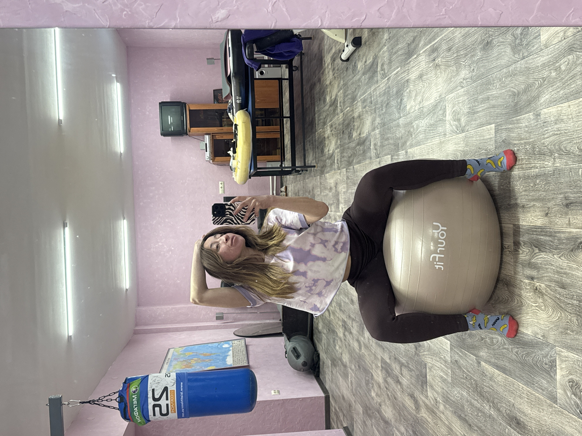 A young woman is lying on a beige exercise ball in what appears to be a home gym. She is wearing dark-colored leggings, a light-colored tie-dye t-shirt, and socks patterned with bananas. She is holding a phone in her hand, seemingly taking a selfie in front of a mirror. The room is illuminated with long, white tube lights. The walls are a light shade of pink. Various gym equipment is visible, including a blue punching bag hanging from a metal frame, an exercise bike, and a floor mat. A map is attached to the wall. The photograph appears to be taken in Прикамский, Russia.