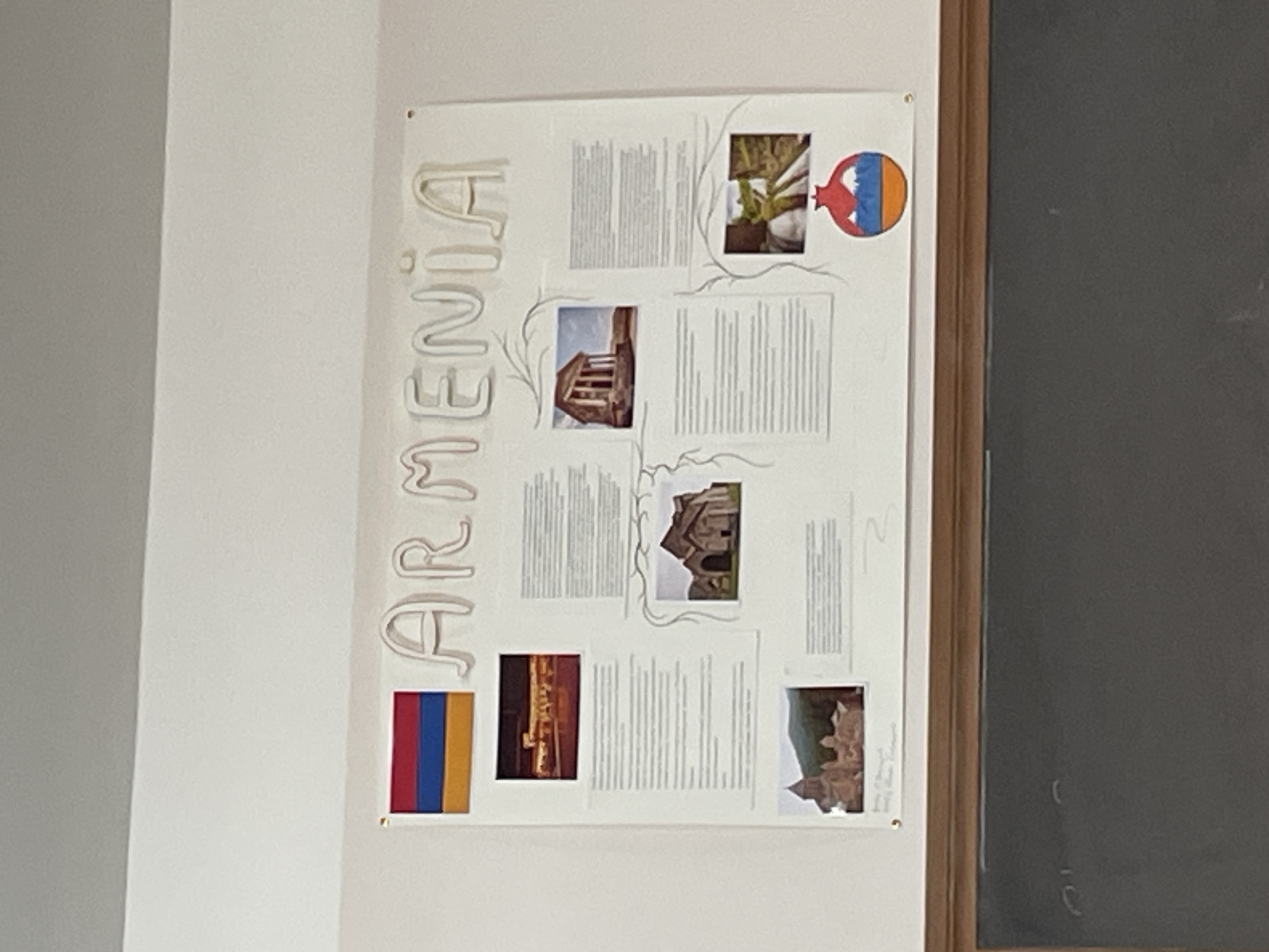 The image shows a poster about Armenia hanging on a wall. The poster has "ARMENIA" written vertically on the left side. It includes the Armenian flag, images of Armenian architecture, a pomegranate with a map, and textual descriptions. The poster appears to be attached to a wall that is adjacent to a dark gray wall, possibly a chalkboard. The location in Yerevan, Armenia is not depicted in the image.
