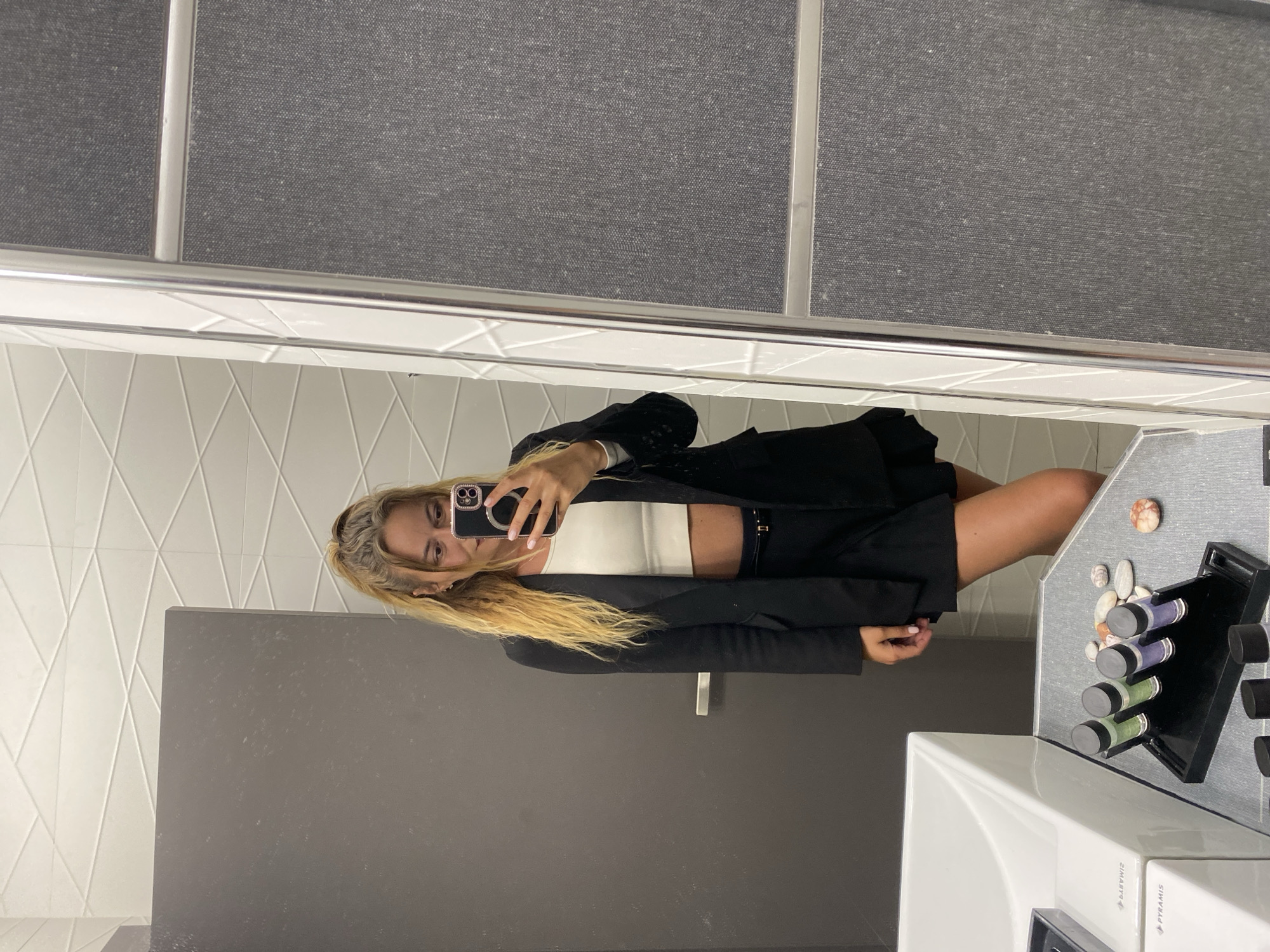 A young woman with long blonde hair is taking a selfie in a bathroom. She is wearing a white crop top, a black blazer, and a black skirt or shorts. Her phone is visible in her hand.
The bathroom has white diamond-patterned tiles on the wall and a dark grey/black door. The sink and countertop are white and grey, respectively. On the countertop are various toiletries and small decorative stones. Above the sink is a mirror with a white frame. A section of grey fabric panels is visible above the mirror.
While the location is specified as Ofrynio Beach, Greece, this detail is not discernible from the visual content of the image alone.