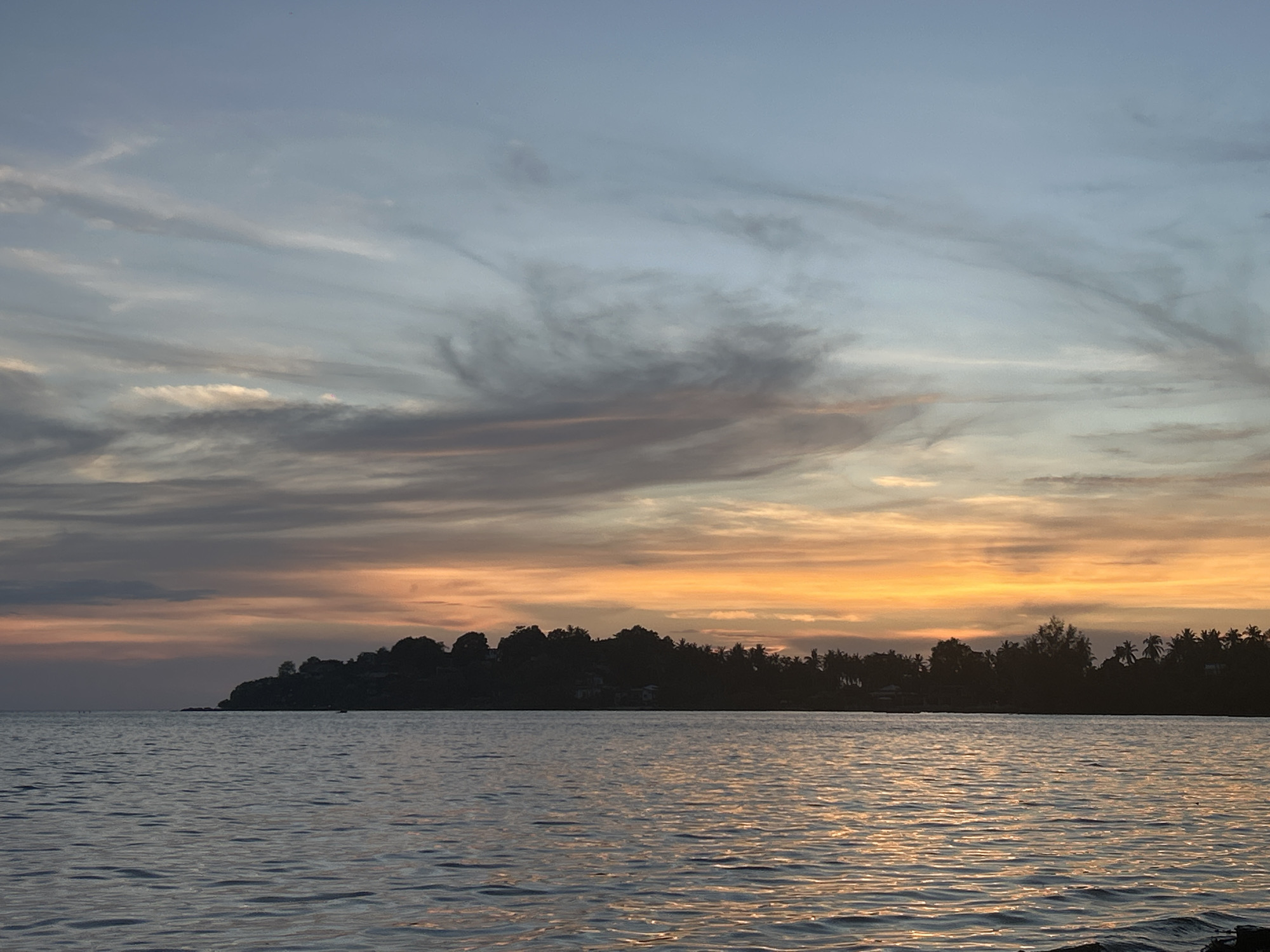 The image shows a sunset scene in ตำบลเกาะพะงัน, Thailand. In the foreground is the ocean with gentle waves reflecting the orange sunlight. A dark silhouette of landmass with trees is in the middle ground. The sky is a mix of blue, gray, orange, and yellow hues with streaks of clouds.