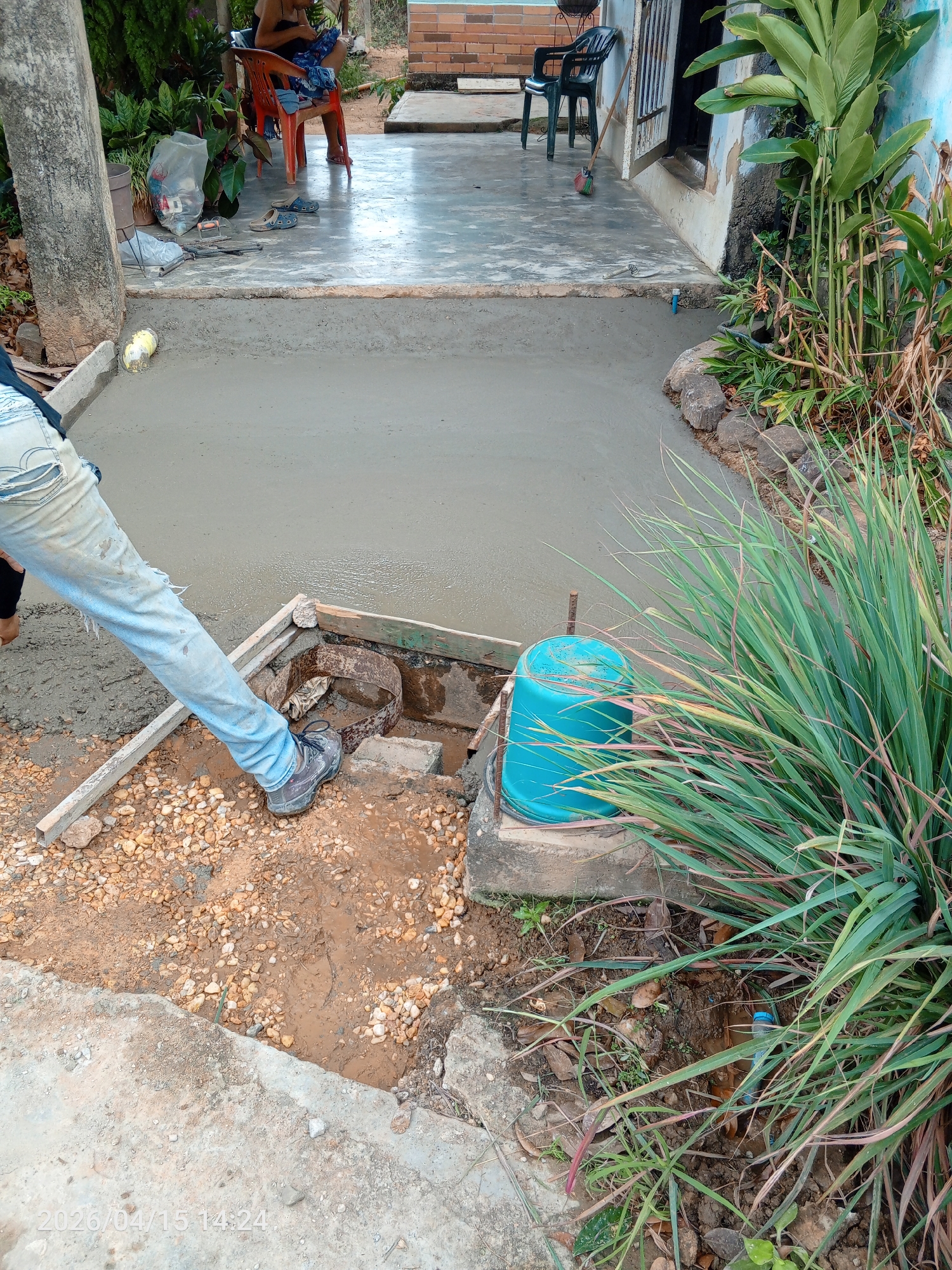 A concrete slab is being poured in an outdoor area in Tinaquillo, Venezuela, as indicated by the timestamp in the image. A person in ripped jeans and dark shoes stands partially in the frame. Concrete is being poured into a section bordered by wooden forms. A circular metal structure with a cylindrical pipe is near the edge of the new slab. A blue bucket sits beside tall grass. There's an existing concrete slab visible in the background and a person sitting in a red chair. A black plastic chair is visible near a building.