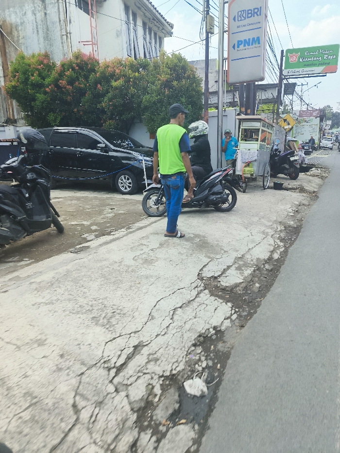 The image shows a street scene in Ngamprah, Indonesia, located at coordinates -6.864 latitude and 107.504 longitude. A man wearing a green vest and blue jeans stands on the sidewalk, looking at a motorcycle parked in front of him. The motorcycle is black with a blue seat and a blue license plate. The sidewalk is cracked and uneven. There is a black car parked in the background. There are also two other people standing in the background, and a food cart. The street is paved, and there are cars and motorcycles parked on the side of the street. The environment is urban, with buildings and trees in the background.