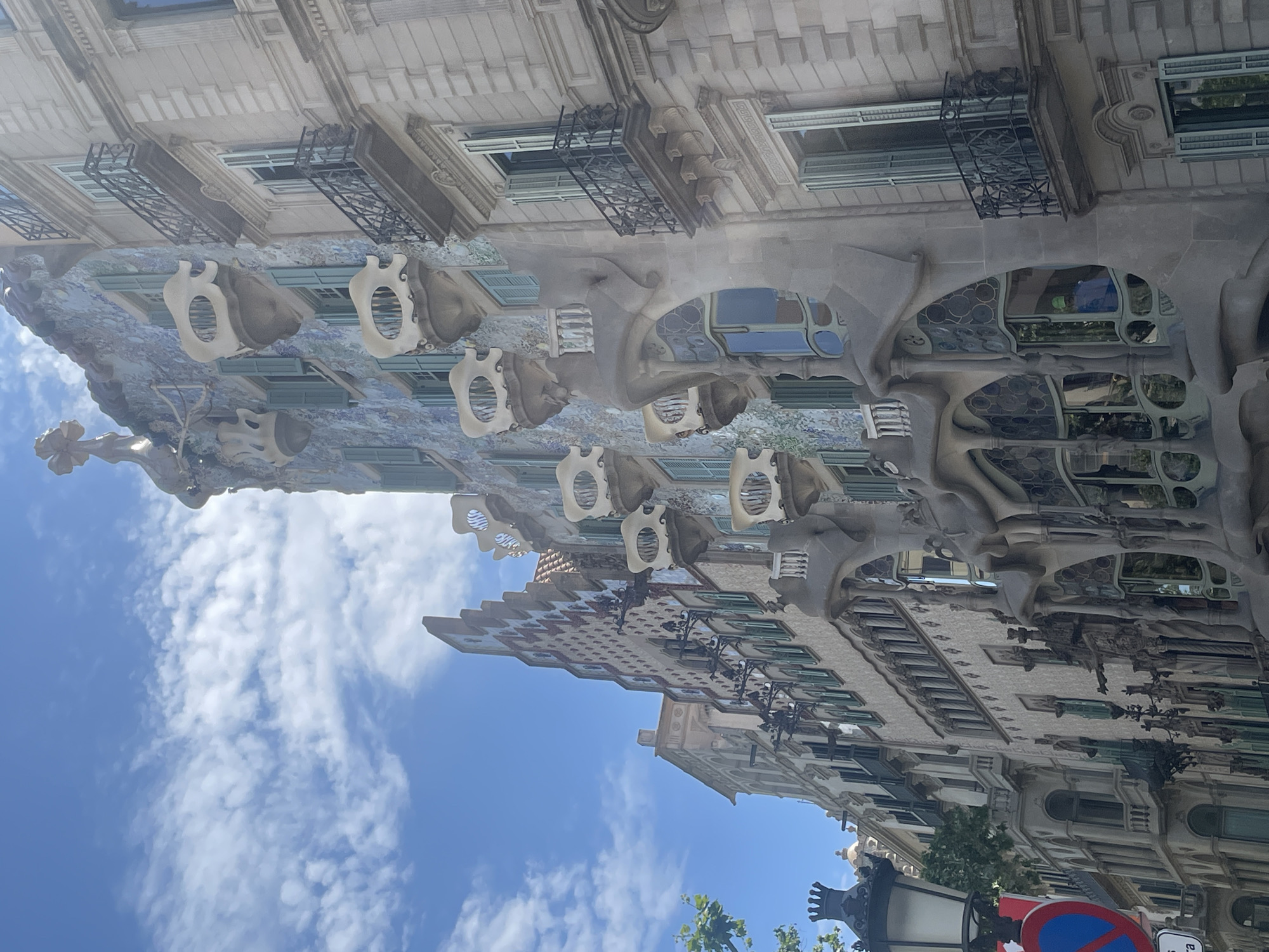 The image captures the iconic facade of Casa Batlló, an architectural masterpiece by Antoni Gaudí, under a bright blue sky with scattered white clouds. The photograph is taken from a street-level perspective, looking up at the building.
The central subject is the highly distinctive and organic architecture of Casa Batlló, which dominates the left side of the frame. Its facade is characterized by flowing, wave-like lines and an iridescent mosaic of blue, green, and beige tiles, giving it a shimmering, aquatic appearance. Protruding from the facade are several pale, bone-like or skull-shaped balconies with irregular openings. The windows are also uniquely shaped, ranging from oval to wavy, some featuring colorful stained-glass effects. Towards the top of the building (visible on the upper left when correctly oriented), the roofline resembles the arched back of a dragon, covered in colorful ceramic scales, complete with a distinctive turret-like structure often interpreted as Saint George's spear.
To the right of Casa Batlló, a portion of an adjacent, more conventionally designed building is visible, featuring standard windows and balconies, contrasting with Gaudí's whimsical design.
The bright sunlight suggests it is daytime, likely midday, with excellent visibility. The clear blue sky and fluffy clouds indicate pleasant weather conditions. No specific human activity is visible, making the focus solely on the architectural forms.
In the bottom right corner, parts of typical urban street furniture are present: a dark street lamp and a blue circular road sign with a red diagonal line, indicating "No Stopping or Standing." Some green foliage from trees can also be seen peeking into the frame from the bottom. There is no other legible text visible, though a small, indistinct logo might be present on the neighboring building to the right. The overall impression is one of architectural wonder and artistic innovation, set against a vibrant urban backdrop.