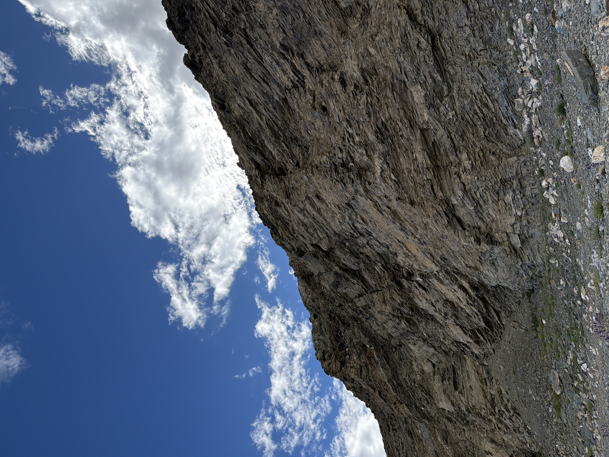 A low-angle, full shot captures a steep, rocky mountainside against a bright blue sky with scattered white clouds. The mountain, predominantly composed of layered, dark brown and gray rock, dominates the right and bottom portions of the frame, sloping upwards towards the top right corner. Its surface is textured with numerous striations and fissures, indicating a geological formation. The base of the mountain is strewn with rocks of various sizes, ranging from pebbles to larger boulders, interspersed with patches of sparse green vegetation and a few small purple flowers. The sky, a vivid blue, fills the upper left portion of the image. White, fluffy clouds drift across the sky, some appearing wispy and thin, while others are more substantial and illuminated by the sun, particularly near the top edge of the mountain. There are no people, animals, or man-made structures visible in the image. The scene appears to be a natural landscape in a mountainous region.
