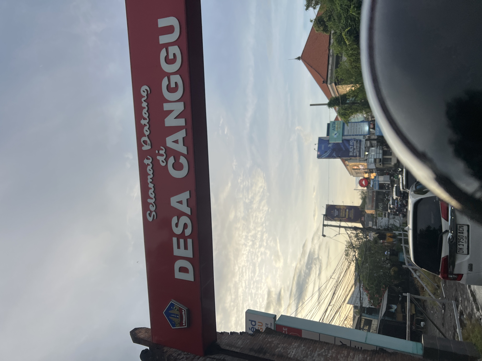 The image shows a street scene in Desa Canggu, North Kuta, Indonesia, as indicated by the "Selamat Datang di DESA CANGGU" sign in the foreground. The sign is red with white lettering. The view is partially obstructed by a large, dark, curved object, potentially a part of a vehicle.
In the background, there is a busy street with various vehicles, including cars and motorcycles. Power lines stretch across the sky. Buildings and billboards are visible along the street. A white car, with the license plate "DK 1750 ACM," is located to the right side of the scene, slightly ahead of the viewer's position. The license plate also shows "09-28". The weather appears bright with some clouds in the sky.