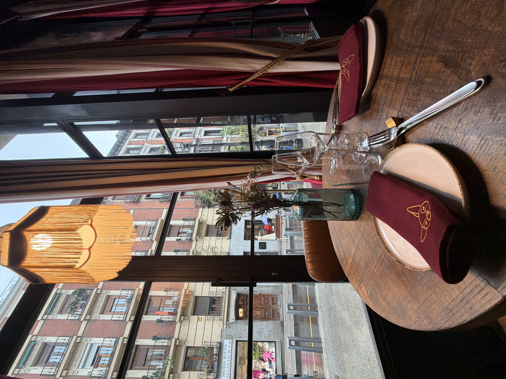 Here is a description of the image:
This interior shot, taken at table level, presents a still life that suggests a restaurant setting, with a view of Madrid's urban architecture through a window. The focal point is a round wooden table set for dining. A fork and knife are neatly placed beside a plate with a folded maroon napkin, embroidered with a stylized dog design. A glass of water sits behind the plate, and a small glass vase containing dried flowers acts as a centerpiece. The table setting is repeated on the other side of the image.
Through the window, a typical Madrid street scene is visible, dominated by apartment buildings with brick facades and balconies. Pedestrians and perhaps storefronts are implied, adding depth to the view. The architecture is indicative of the city's historic districts.
The image's composition balances the indoor dining arrangement with the outdoor cityscape, implying a location within a Madrid restaurant or bar where the interior design emphasizes natural light and urban views.
