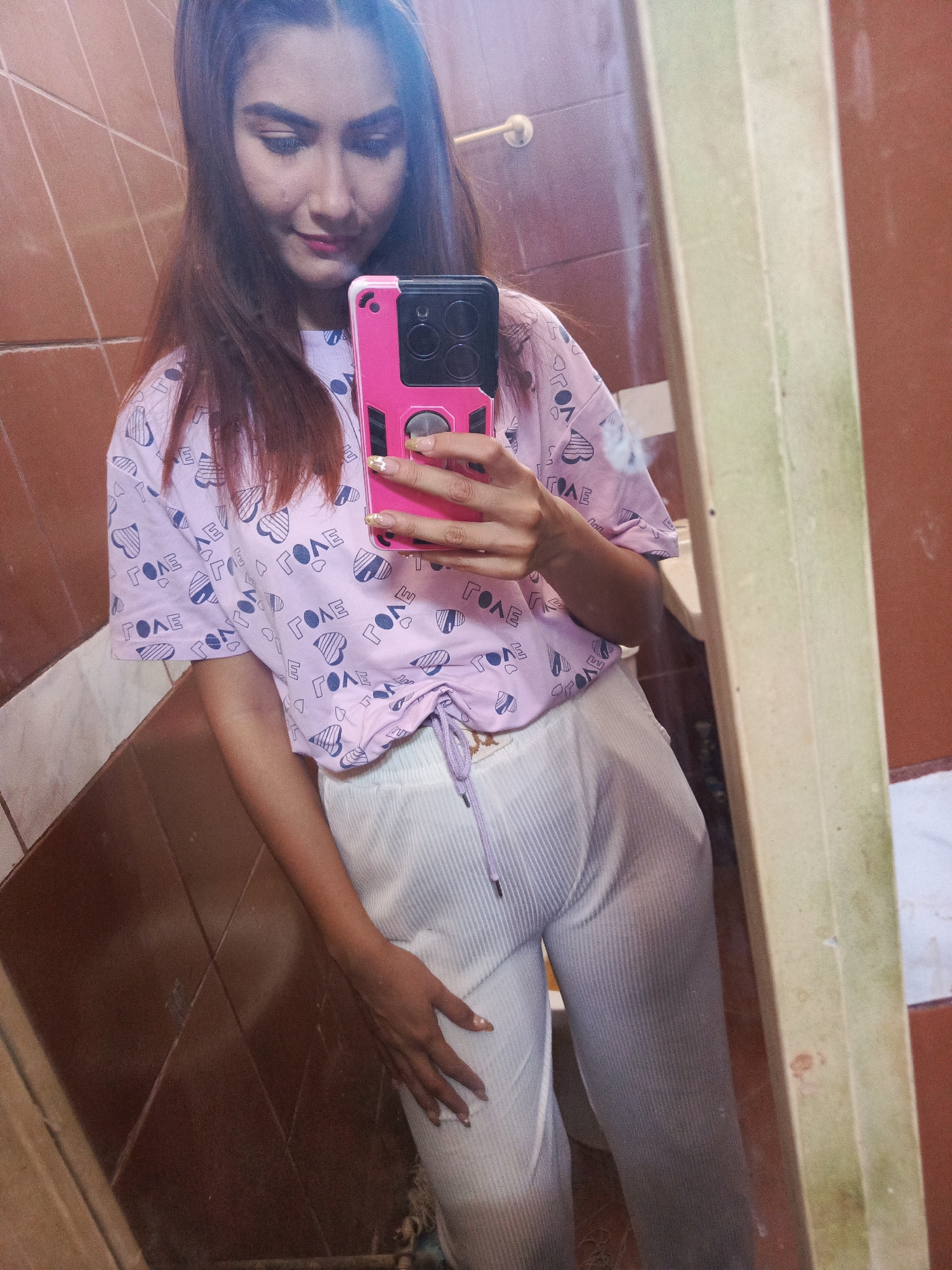 A young woman is taking a mirror selfie in what appears to be a bathroom. She is wearing a light purple t-shirt patterned with the word "LOVE" and hearts, and light-colored, pleated pants. She is holding a pink and black phone in her right hand. The bathroom has brown tiled walls and a partially visible sink. The reflection in the mirror shows a brown door. The exact location in Juan Griego, Venezuela is not discernible from the image.