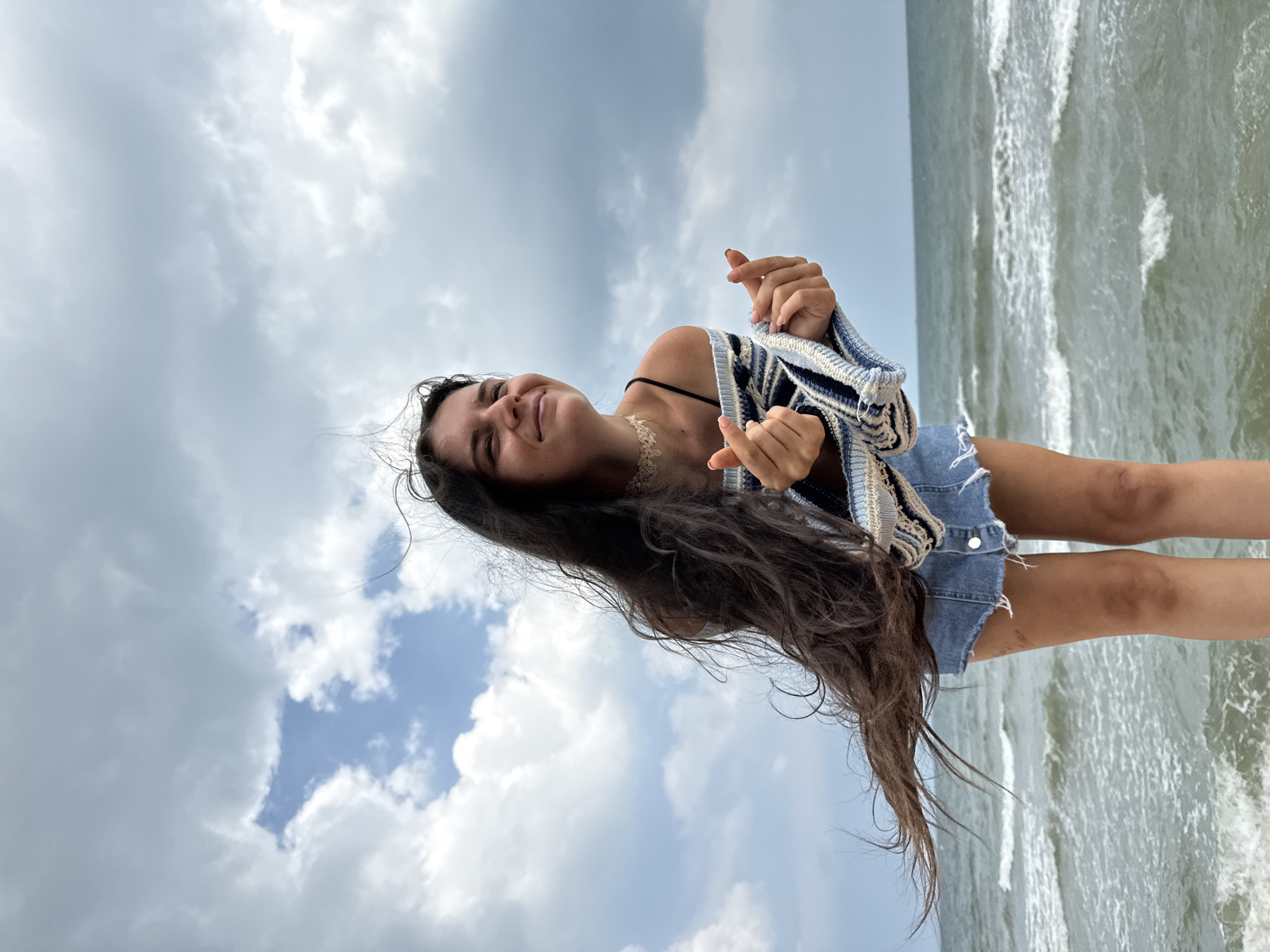A young woman with long, dark hair stands on a rocky outcrop overlooking the ocean under a cloudy sky. She is wearing a striped sweater draped off her shoulders and denim shorts. Her hands are held up in front of her, forming finger hearts. The waves are crashing against the shore behind her. The location is Phu Thuy Ward, Vietnam.