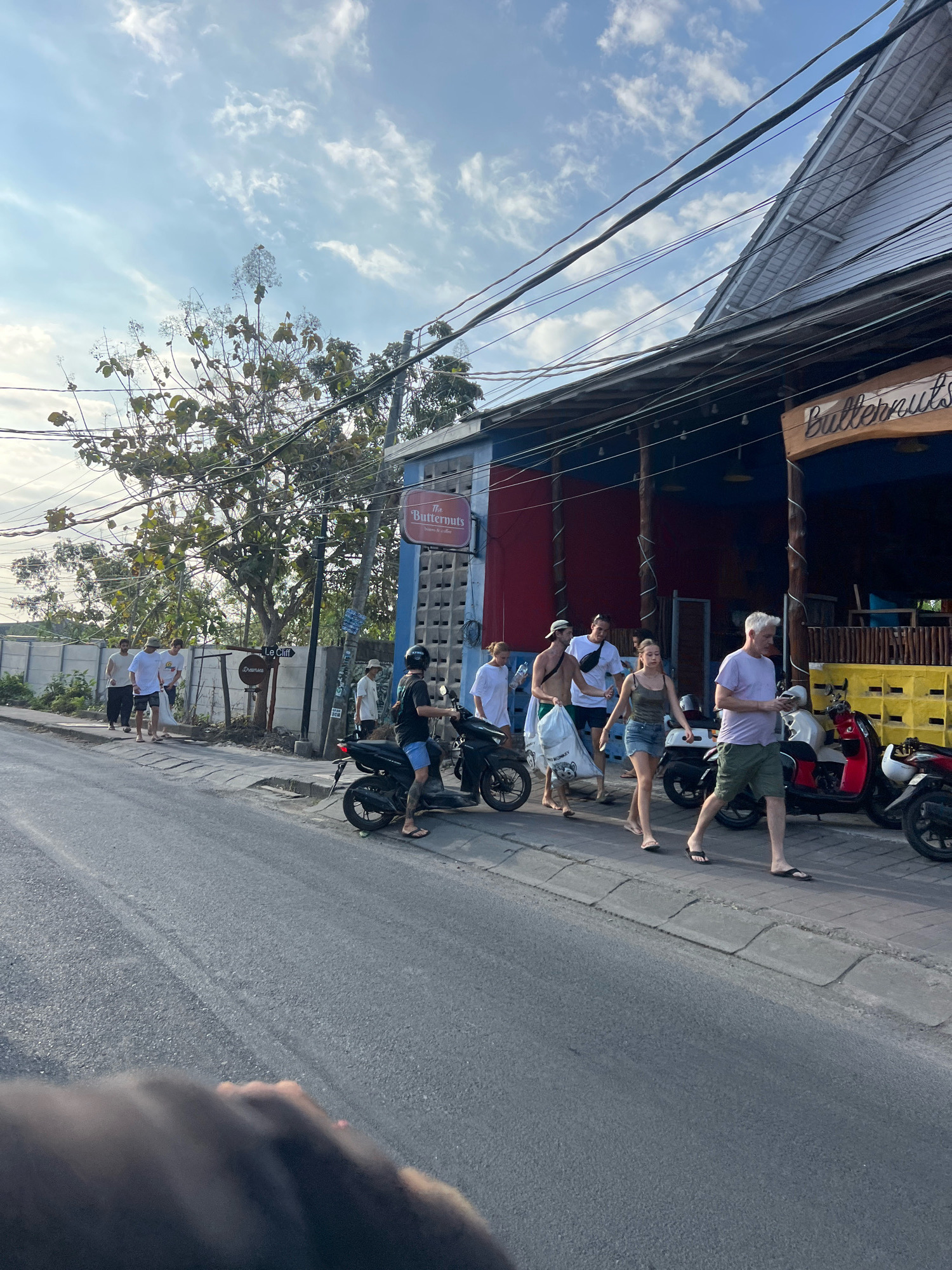 In Kuta Selatan, Indonesia, a group of people walk along a paved sidewalk next to a street. The street is lined with buildings, one of which is a cafe named "The Buttercups." Several motorbikes are parked on the side of the road. A tree with sparse foliage grows on the left side of the image. The sky is partly cloudy. The overall scene depicts a casual street view with pedestrian and vehicle activity.