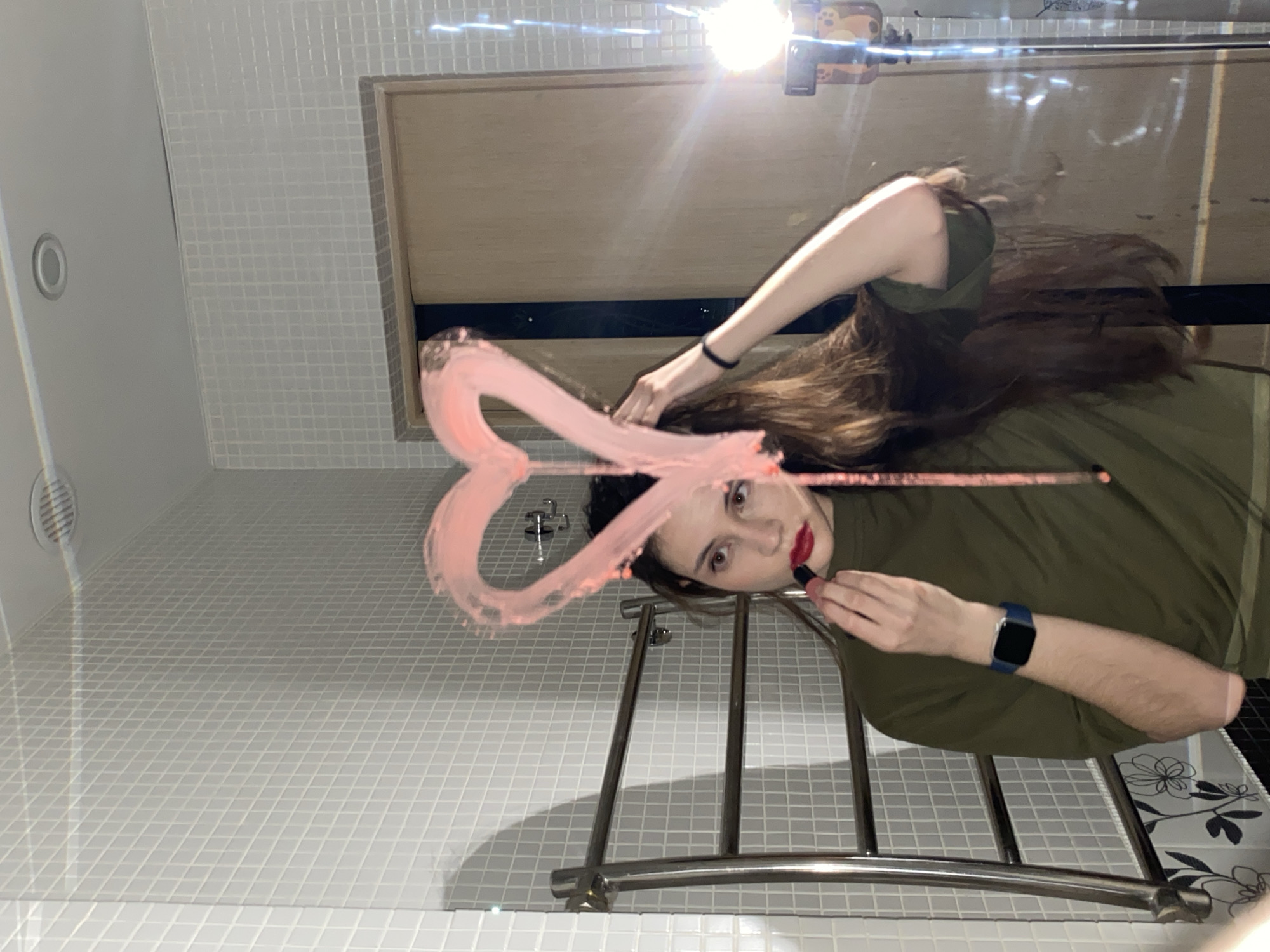 This image captures a close-up, reflective view of a person applying makeup in what appears to be a bathroom in Votkinsk, Russia. The photo is taken from a low angle, showing the subject's reflection in a mirror.
The main subject is a young woman with long, dark brown hair, wearing an olive-green t-shirt. She is looking directly into the mirror, holding a red lipstick tube to her lips. She has already applied some red lipstick. On her left wrist, she wears a blue smartwatch.
A prominent detail in the reflection is a large, pink heart shape drawn with lipstick on the mirror surface, positioned to the left of the woman's face. The bathroom setting is indicated by the white, square-tiled walls and floor visible in the reflection. A silver-colored metal towel rack or similar fixture with three horizontal bars is reflected in the lower central part of the image.
In the upper right corner of the reflection, a bright light source creates a strong glare, and a smartphone in a patterned, light brown case (possibly with paw prints) is visible, likely the device used to capture the image. Other reflected elements include a circular recessed light fixture and a rectangular vent on what appears to be the ceiling or upper wall. The overall lighting is bright, suggesting indoor artificial light. The woman's expression is focused on her activity. No visible text is present.