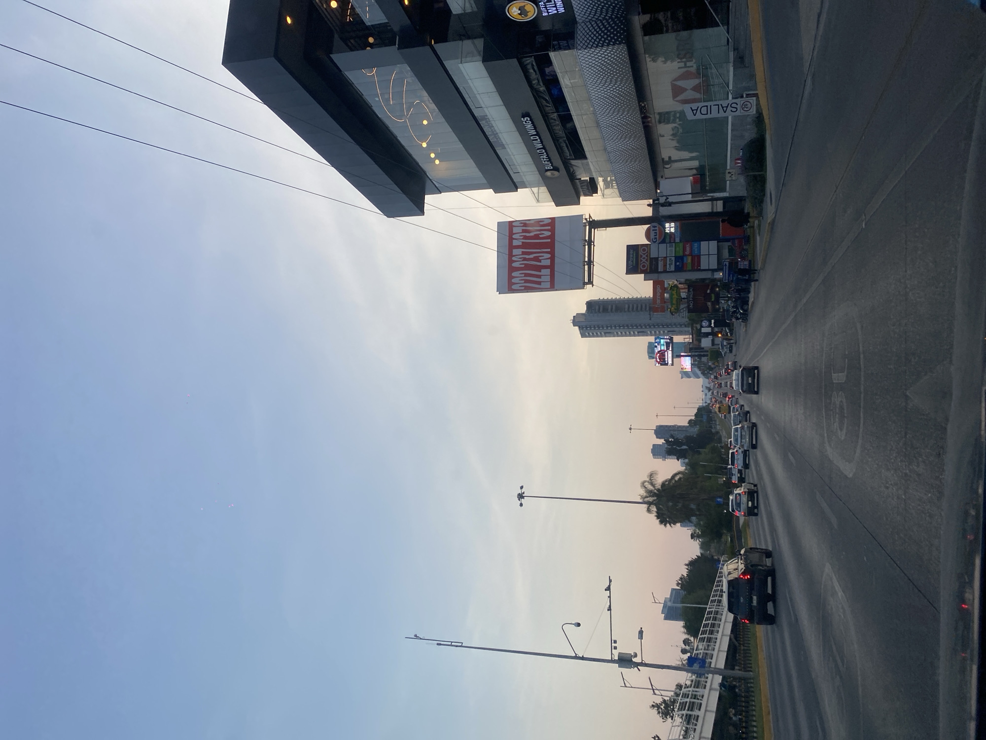 The image depicts a city street at dusk, featuring a multi-lane road filled with cars traveling away from the viewer. On the right side of the road is a sidewalk with a bridge. Above the road and sidewalk are street lights. The left side of the image shows a large commercial building with businesses such as Buffalo Wild Wings and HSBC. There are also signs for OXXO and Gulf gas stations. A large sign with the number 222 237 7378 is visible. The sky is light blue, fading into a reddish-orange hue.