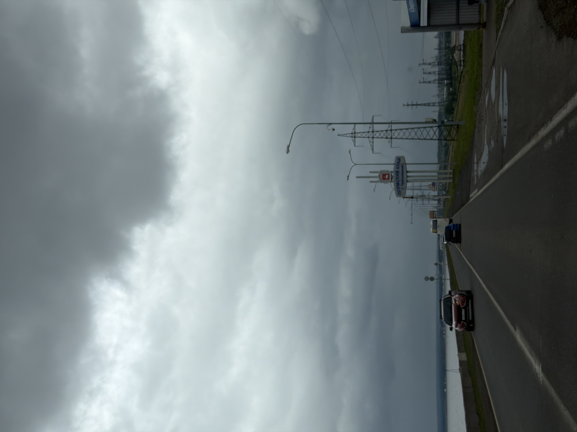 The image depicts a road scene under an entirely overcast, grey sky, suggesting dim and diffused lighting conditions. The specific location is identified as Perm Krai, near Chaykovsky city, Russia, based on a prominent road sign.
The foreground features a two-lane asphalt road, with white lane markings, extending into the distant horizon. On the left side of the road, a wide body of water, likely a river or reservoir, is visible beyond a low concrete embankment and a strip of green vegetation.
Vehicles are present on the road. A red SUV, appearing to be a Lada XRAY Cross, is observed in the left lane, driving towards the camera. Behind it, a smaller blue car is also traveling in the same direction. On the right side of the road, a white bus is partially visible, facing away from the camera, positioned near a road sign.
The right side of the road includes a paved shoulder with several visible puddles, indicating recent precipitation. Further right, a grassy verge is present. Multiple streetlights and utility poles equipped with overhead wires line this side of the road. A large informational road sign, supported by multiple vertical poles and topped with a red insignia, displays Cyrillic text that translates to "PERMSKIY KRAY" (Perm Krai) and "G. CHAYKOVSKY" (Chaykovsky city). In the mid-ground and distant background on the right, numerous lattice-steel electricity transmission towers (pylons) are visible, along with industrial structures on the far horizon.