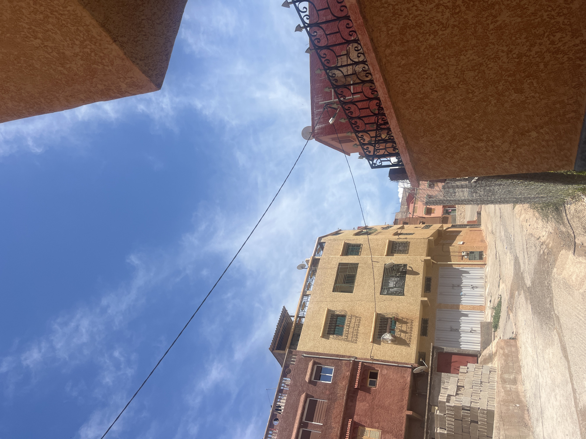 The image depicts a street view in Ouaouizert, Morocco. The perspective is looking upwards through a narrow alleyway between buildings. The sky is blue with scattered clouds. Buildings on either side are constructed of brown and yellow stucco with ornate wrought iron balconies. A power line stretches across the sky. At ground level, the street is paved and a stack of grey blocks is visible.