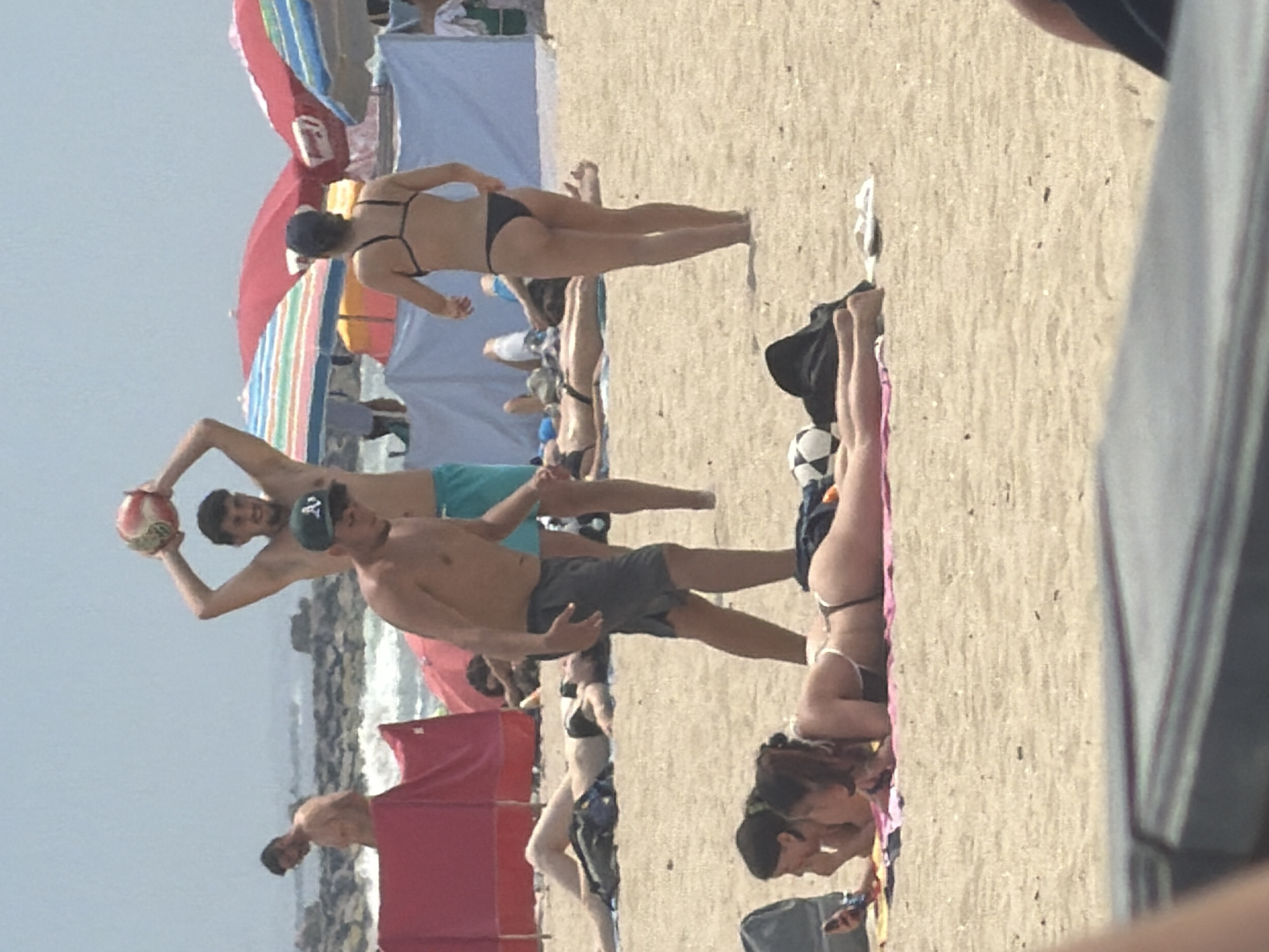This image captures a lively beach scene in Espinho, Portugal, on a sunny day. In the foreground, two women are lounging on the sand, one with her arm extended and the other looking at a phone. Behind them, a group of people are enjoying the beach.
A man in the center is holding a volleyball above his head, as if preparing to serve. To his left, another man is standing and looking towards the sea. A woman in a black bikini is facing away from the camera, with her back to the viewer. In the background, colorful beach umbrellas and tents are visible, along with a glimpse of the ocean and rocky coastline. The sand is light brown and speckled with small shells and debris. The overall atmosphere is one of a typical summer day at the beach, with people relaxing and engaging in recreational activities.