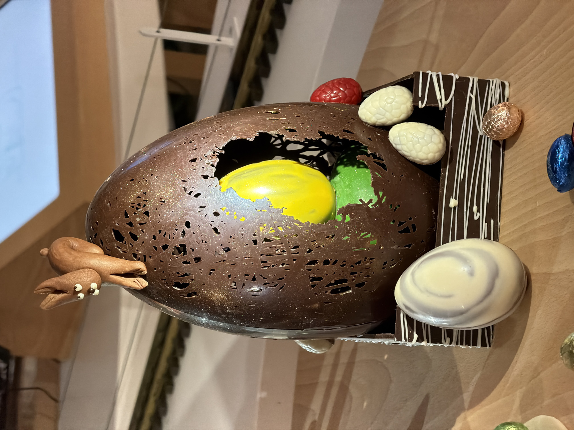 The image shows a large, elaborately decorated chocolate Easter egg display. The egg is mostly brown with a textured, hole-filled surface. Inside, there are yellow and green spheres, likely made of chocolate or candy. A small chocolate rabbit figurine is attached to the side of the egg. The egg is mounted on a dark chocolate stand with white chocolate drizzle decoration, and several smaller eggs are attached to the stand, including white, marble, and foil-wrapped eggs. A red, berry-shaped candy is also visible on top. The background is a light-colored wooden wall with vertical panels. The display is located at Thierry Mulhaupt, Illkirch-Graffenstaden, France.