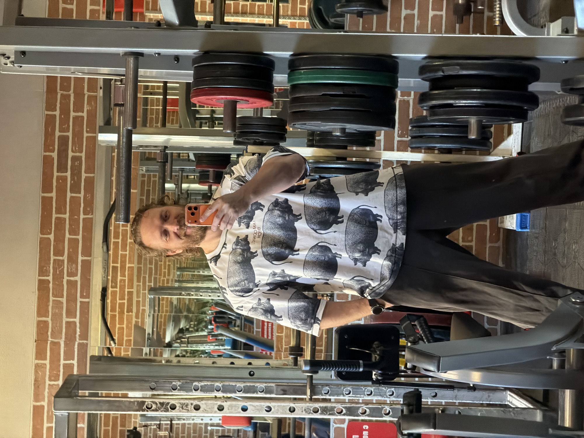 The image displays a mirror selfie of a man inside a gym or weightlifting facility. The man, who has medium-length light brown hair and a beard, is wearing a distinctive white t-shirt covered in multiple black and grey pig patterns, along with dark pants and a dark watch on his left wrist. He holds an orange-cased smartphone in his right hand, angled to capture his reflection, and has a slight smile on his face.
The setting is characterized by substantial gym equipment. Prominently featured are heavy-duty metal racks or a power cage structure, which store numerous weight plates on various posts. These plates include black, red, and green colored ones, as well as some lighter (possibly white or cream) bumper plates. The entire scene is presented as a reflection in a large mirror, which also reveals a brick wall in the background of the gym, and indistinct reflections of other gym apparatus. The lighting is bright, illuminating the indoor space. A small, partially obscured red sign is visible through the equipment at the bottom of the frame, but any text on it is illegible.