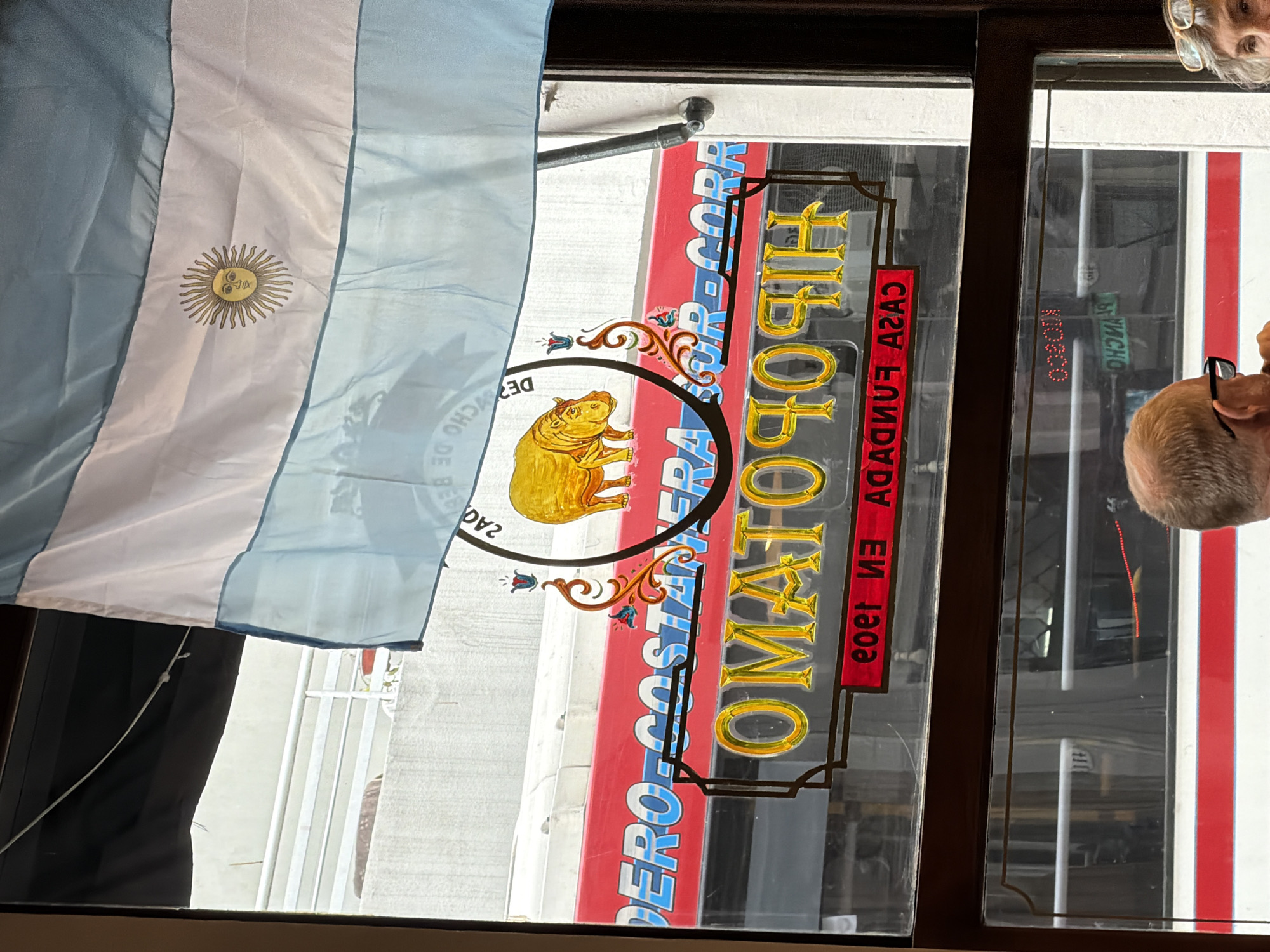 The image shows a window of a business in Buenos Aires, Argentina, partially obscured by an Argentine flag hanging from inside. The sign on the window reads "HIPOPOTAMO CASA FUNDADA EN 1900," with a golden hippopotamus logo. The text "PERO-COSTANERA SUR-CORA" is also visible on the sign. Reflections in the glass show a blurred street scene, and two people are partially visible in the reflections, one with glasses. The scene suggests the entrance of a historical establishment.