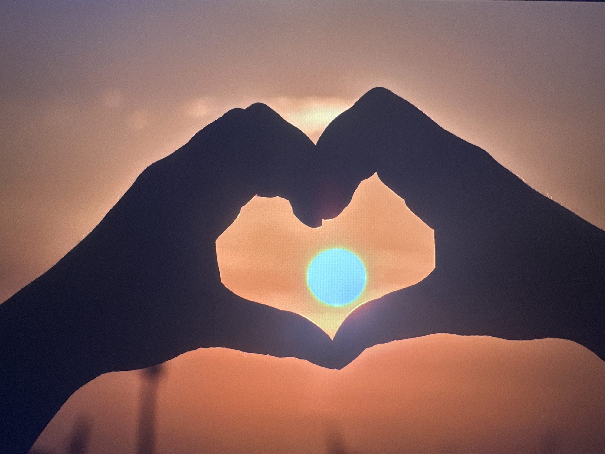 The image is a close-up silhouette of two hands forming a heart shape against a sunset sky. The sun, appearing as a bright, teal-colored circle, is positioned in the center of the heart. The sky transitions from a warm orange hue near the horizon to a cooler, muted purple at the top. The hands are dark silhouettes, with the fingers curved to create the heart shape. The background shows a hazy, slightly textured sky with some indistinct lighter areas, possibly clouds or atmospheric effects. The setting appears to be an outdoor environment, likely a landscape or open area, given the vastness of the sky. There is no visible indication of the city of Bryansk, Russia.