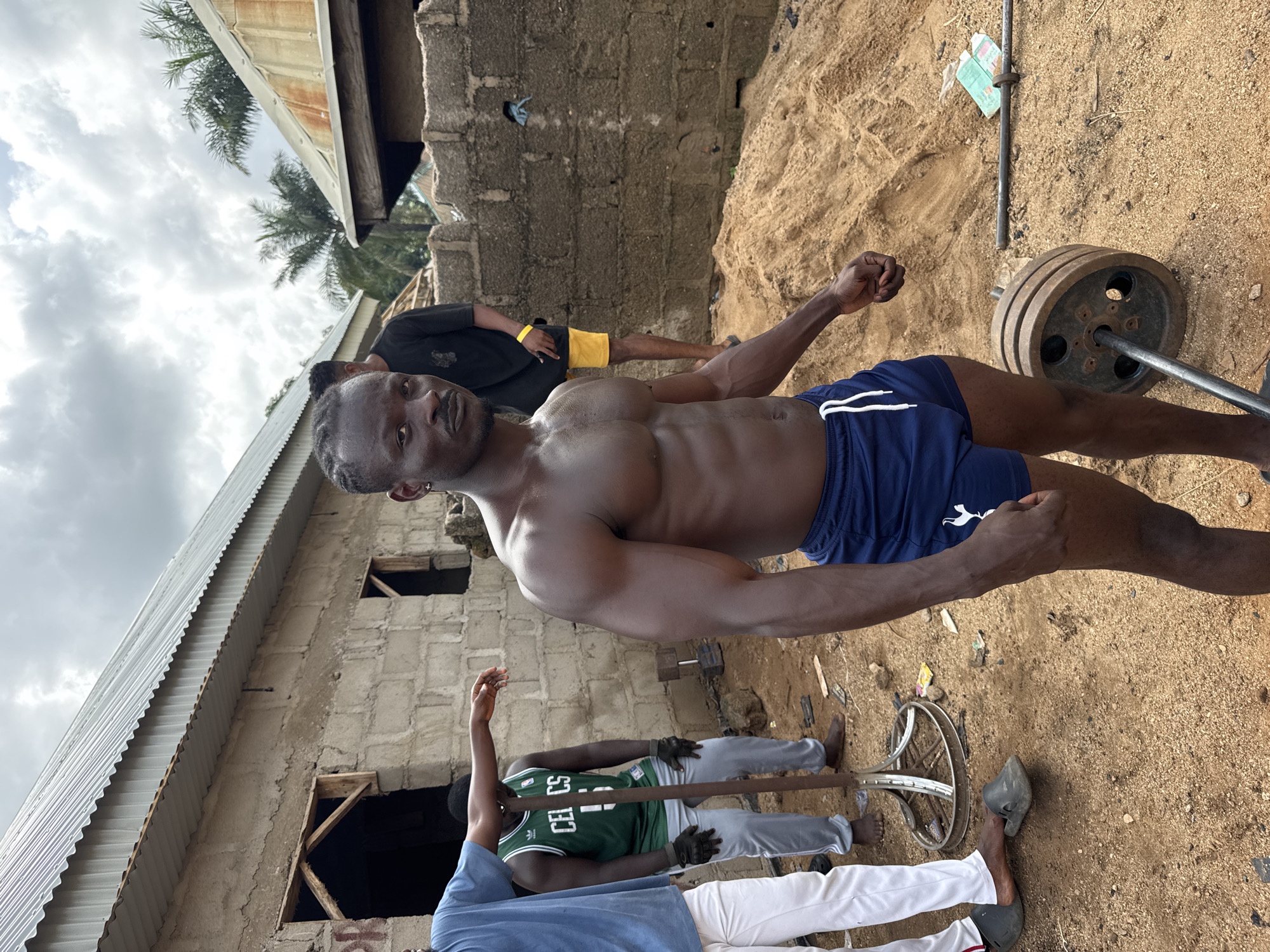 A photograph taken in Akwanga, Nigeria, shows a muscular, shirtless man wearing blue shorts standing outdoors. The environment appears to be a construction site with sand and unfinished buildings. Other individuals are present, including one wearing a green basketball jersey and another with a black shirt and yellow shorts. A barbell with weights is also visible.